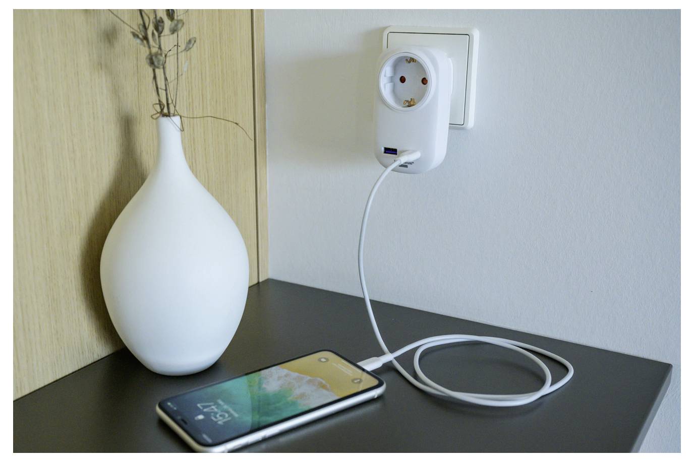 Smartphone wird auf einer Tischplatte neben einer Steckdose aufgeladen, mit einem aufgewickelten weißen Kabel, das eingesteckt ist, und einer weißen Vase im Hintergrund.