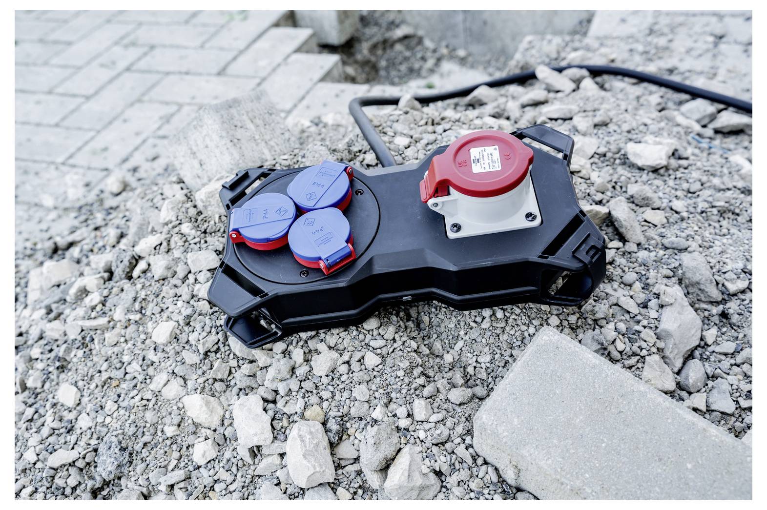 Tragbare Stromverteilungsbox mit roten und blauen Steckdosen liegt auf Kies und Bauschutt neben einer gepflasterten Oberfläche.