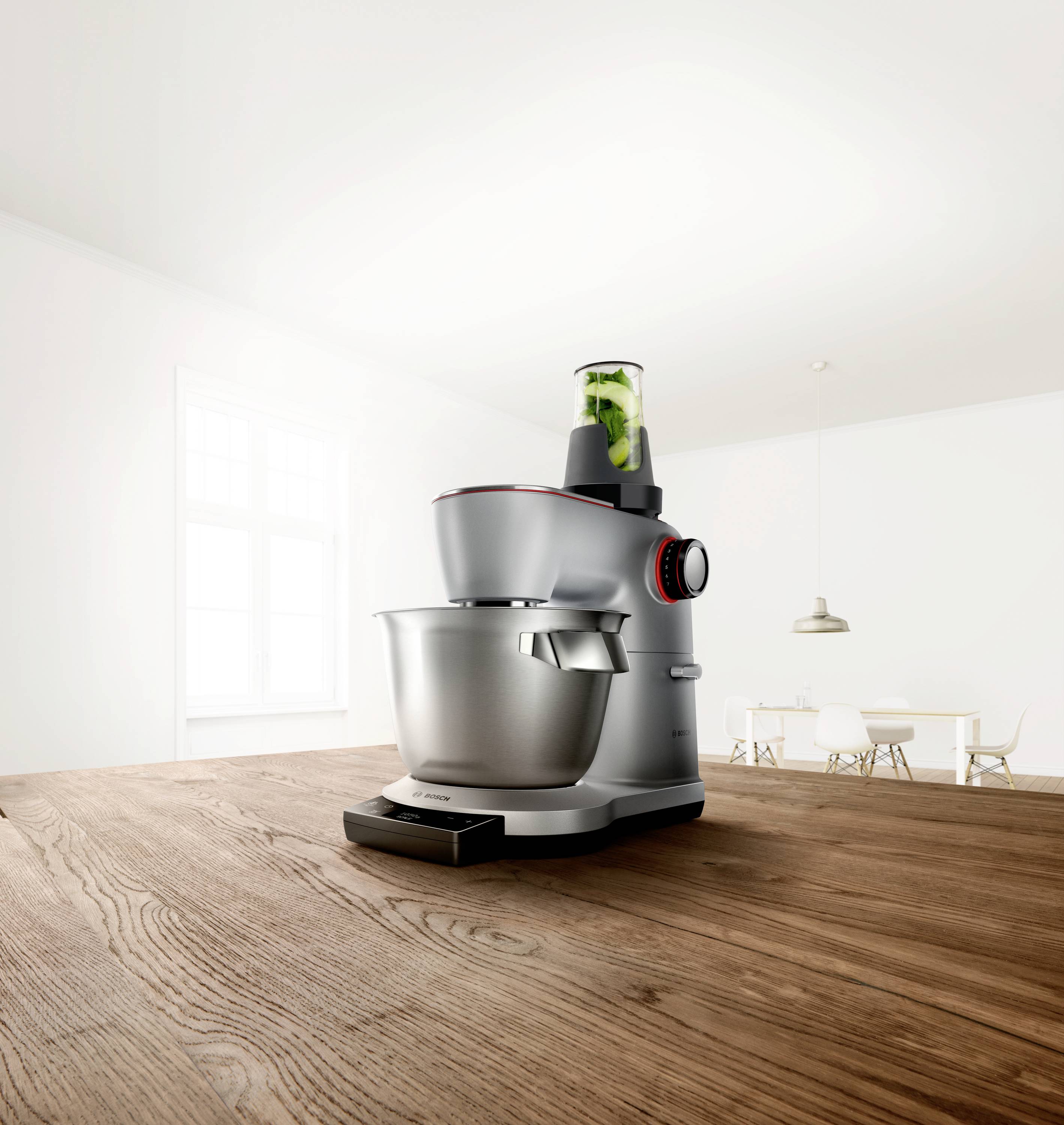 Eine moderne Küchenmaschine mit einer Metallschüssel und einem Mixaufsatz, der grüne Zutaten enthält, steht auf einem Holztisch in einer hellen Küche.