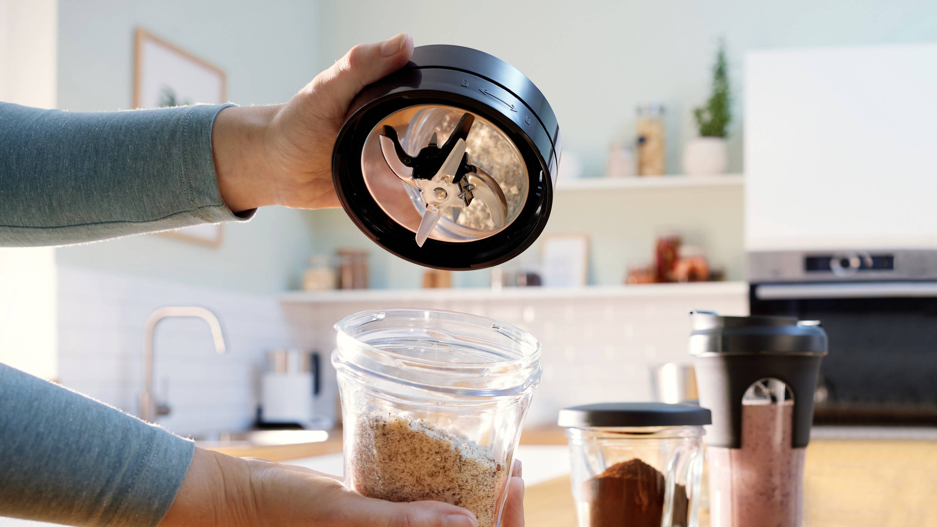 Eine Person hält einen Mixer-Deckel mit Klingen über einen Glaskrug mit gemahlenem Kaffee in einer Küche und bereitet sich darauf vor, zu mixen.