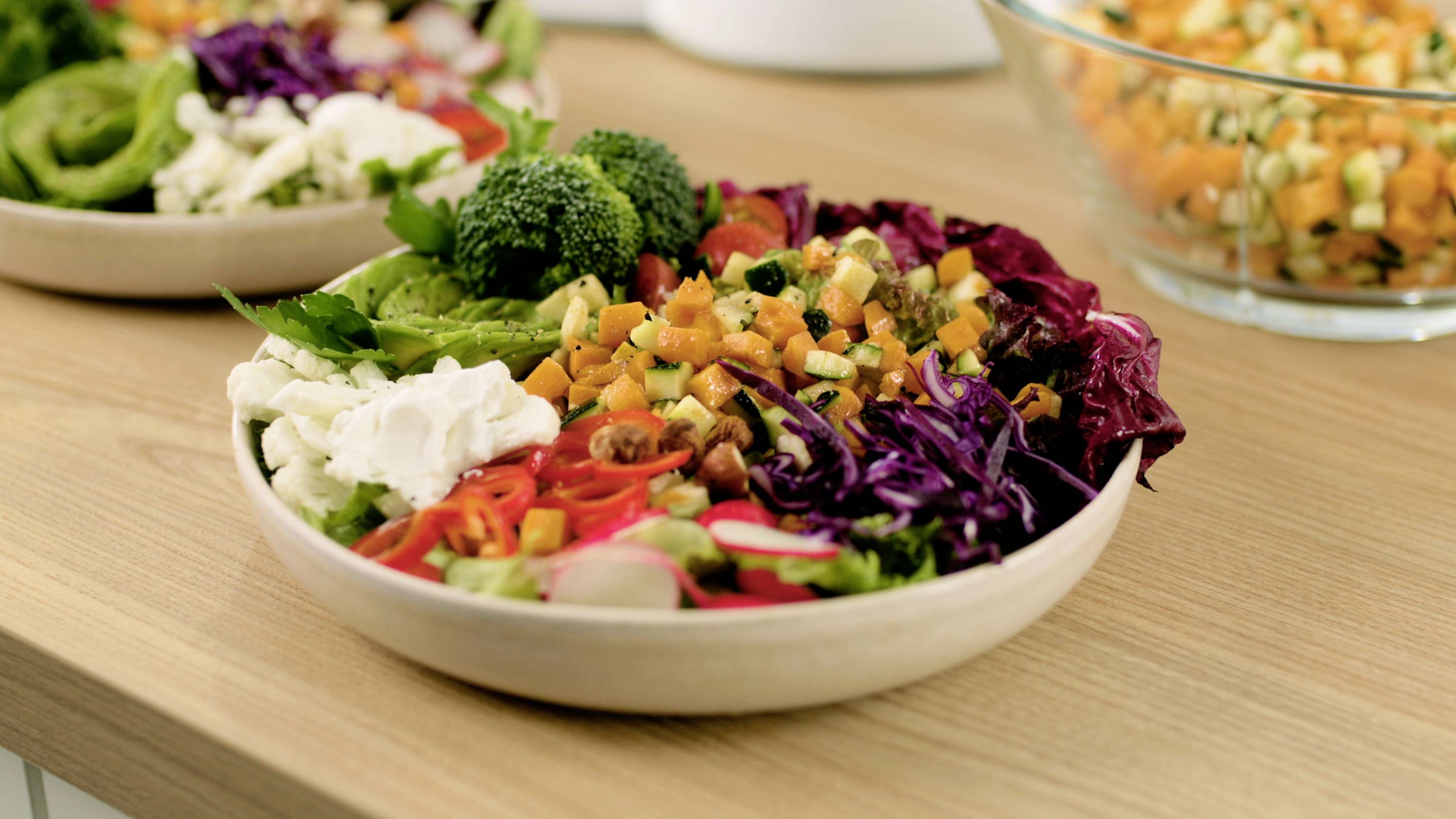 Eine Schüssel bunter gemischter Salat mit Brokkoli, Salat, Radieschen, Rotkohl, gewürfelten orangefarbenen Gemüsestücken und Gurken auf einem Holztisch.