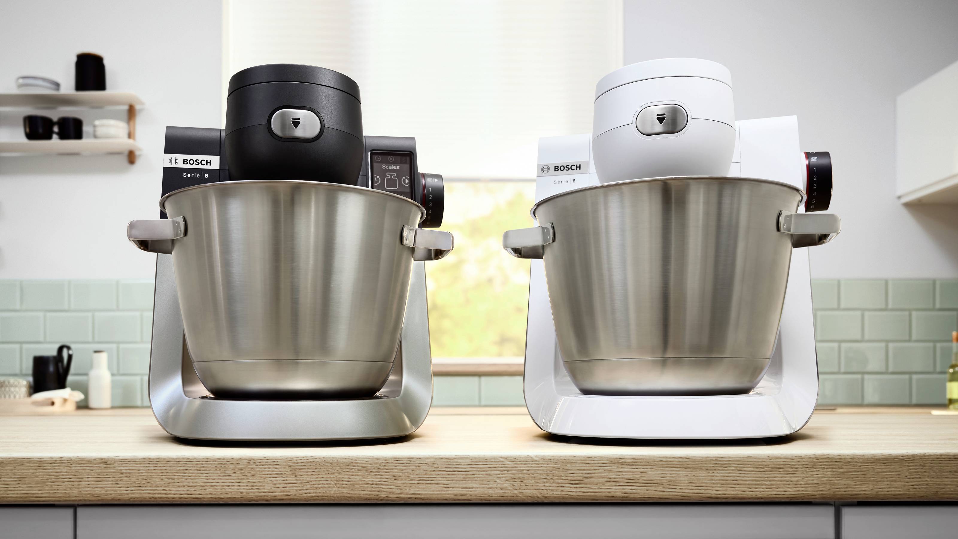 Zwei Bosch Küchenmaschinen, eine in Schwarz und eine in Weiß, stehen auf einer Holzarbeitsplatte in einer Küche mit einem Fenster und gefliester Rückwand.