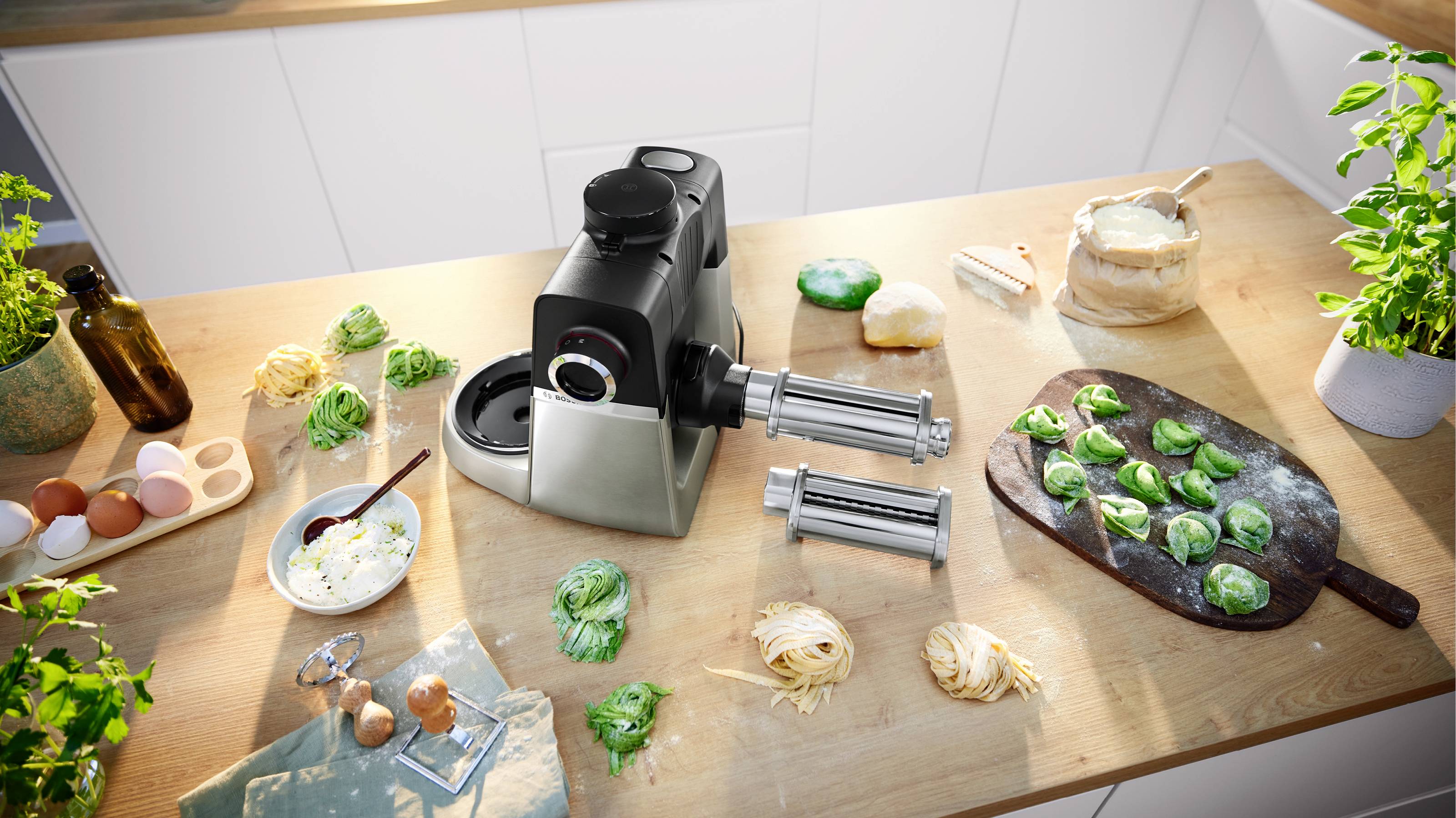 Nudelmaschine auf einer Küchenarbeitsplatte mit frisch gemachten grünen und gelben Nudeln, Eiern und Kräutern, was auf selbstgemachtes Kochen hindeutet.