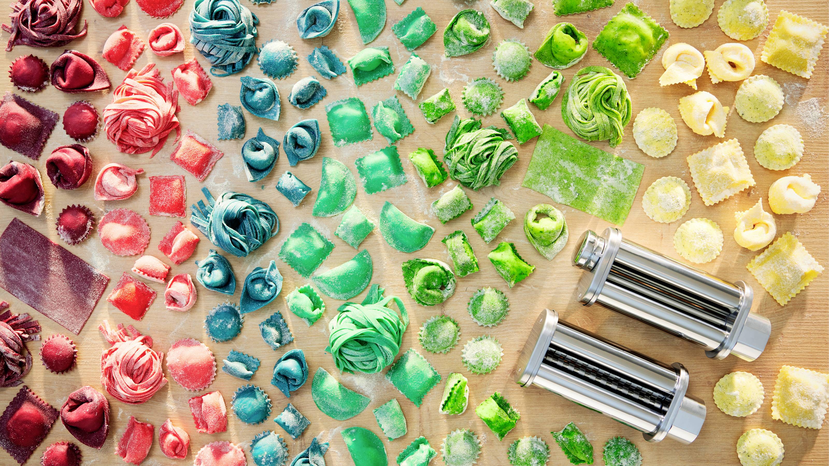 Eine Anordnung von mehrfarbiger Pasta in verschiedenen Formen, darunter Ravioli und Fettuccine, in Abschnitten nach Farbe sortiert, mit zwei Pasta-Schneidern daneben.