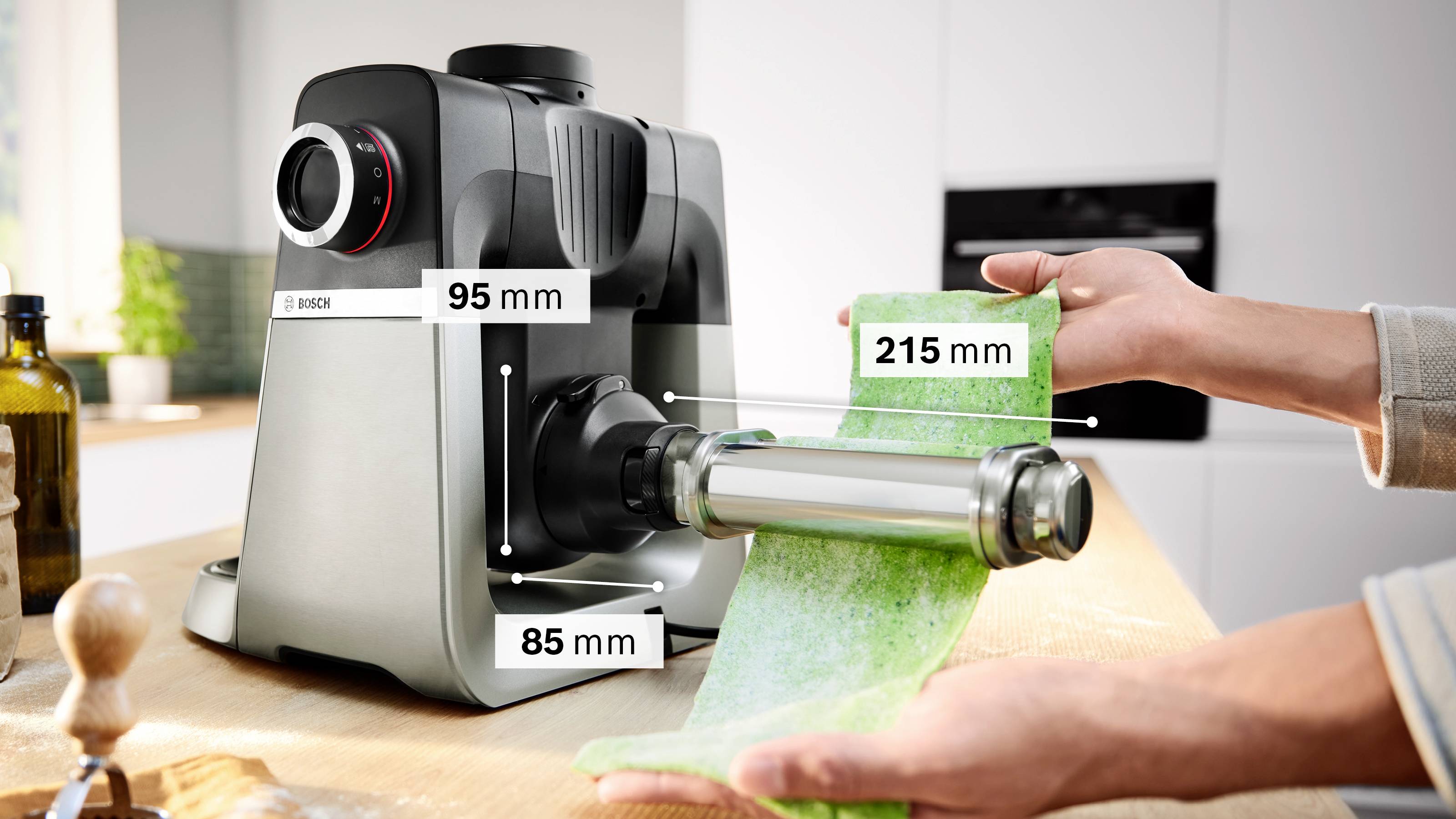 Eine Nudelmaschine ist auf einer Küchenarbeitsplatte in Betrieb, mit einer Grundfläche von 95 mm und 85 mm. Die Breite der Nudelwalze beträgt 215 mm. Zwei Hände führen grünen Nudelteig durch die Maschine.