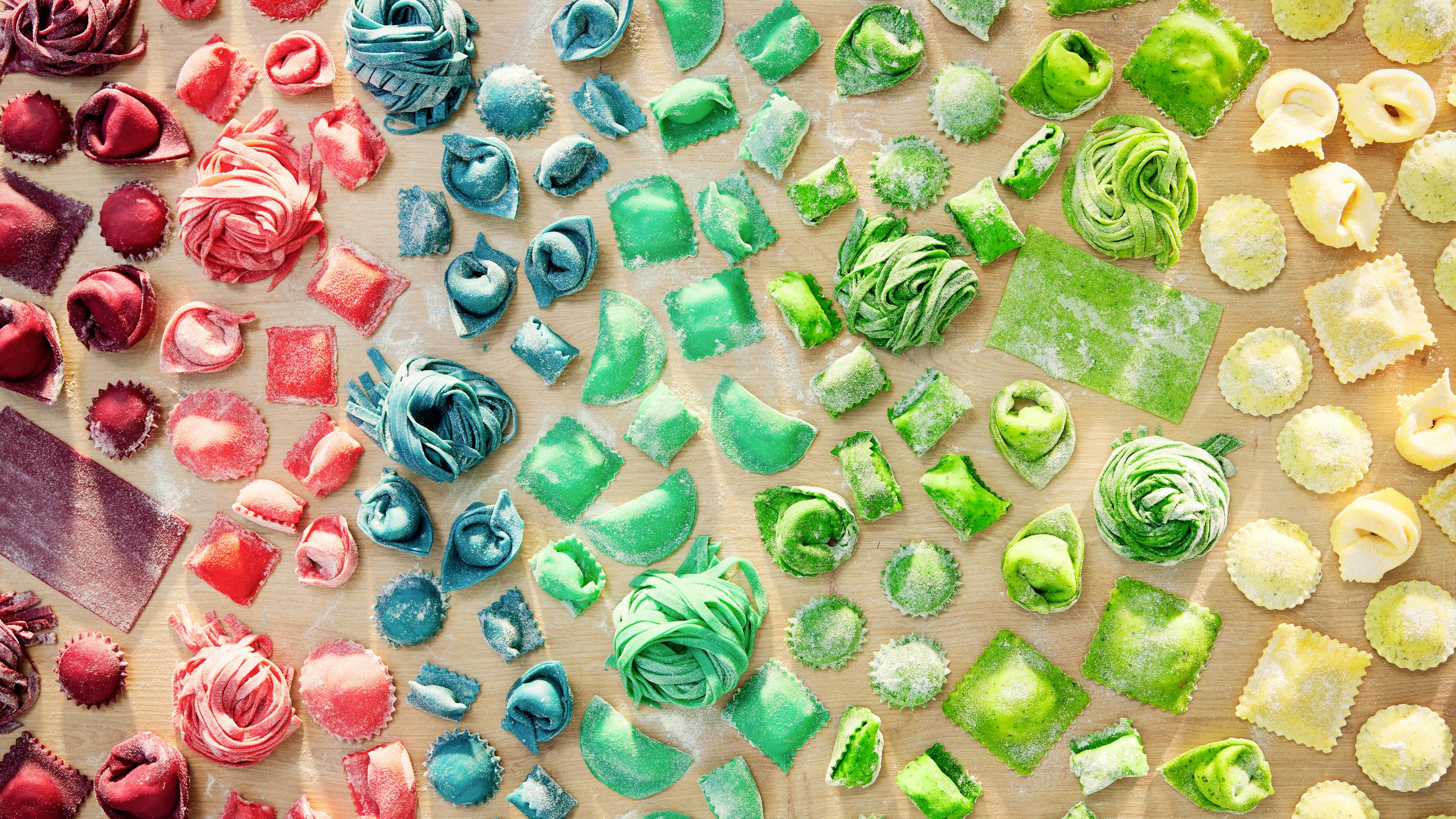 Eine Vielfalt an bunter, handgemachter Pasta, darunter rote, blaue und grüne Nudeln, die in einem Verlaufsmuster auf einer Holzoberfläche angeordnet sind.