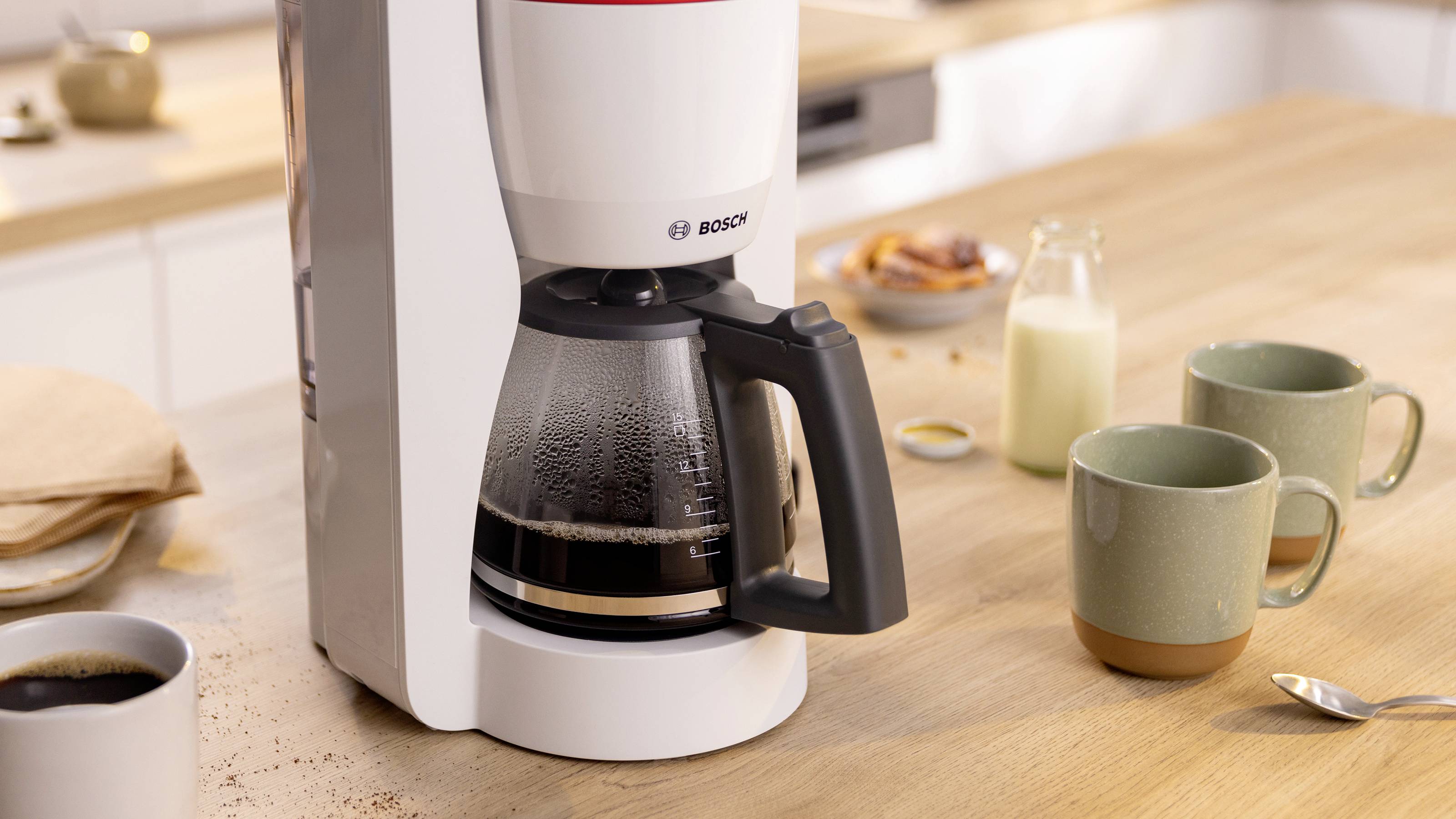 Eine weiße Kaffeemaschine mit einer Glaskanne, gefüllt mit gebrühtem Kaffee, steht auf einer Holztheke. In der Nähe befinden sich zwei Tassen und eine Milchflasche.