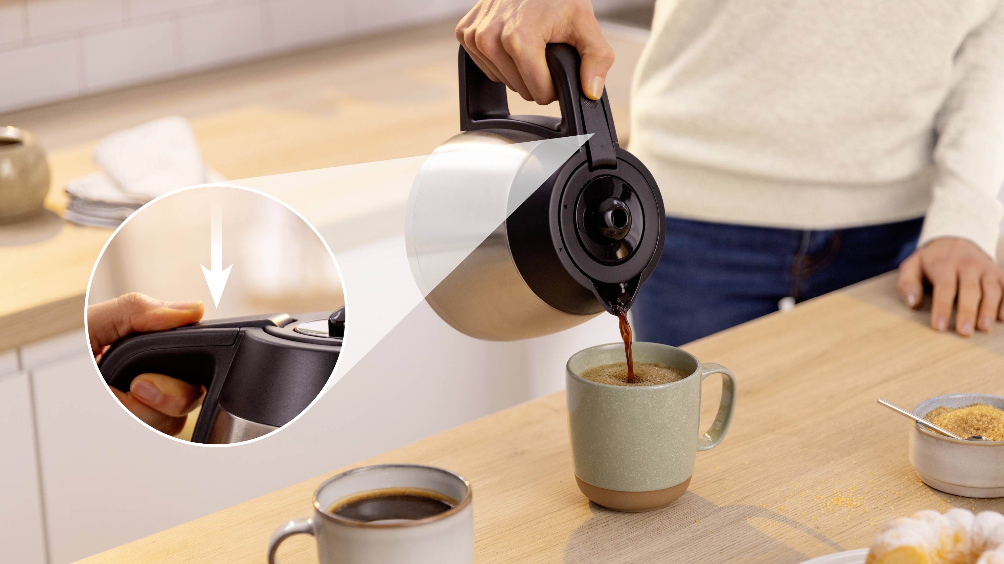 Eine Person gießt Kaffee aus einer Thermoskanne in eine Tasse auf einer Küchenarbeitsplatte. Eine Detailansage zeigt einen Nahaufnahme der offenen Ausgießöffnung.