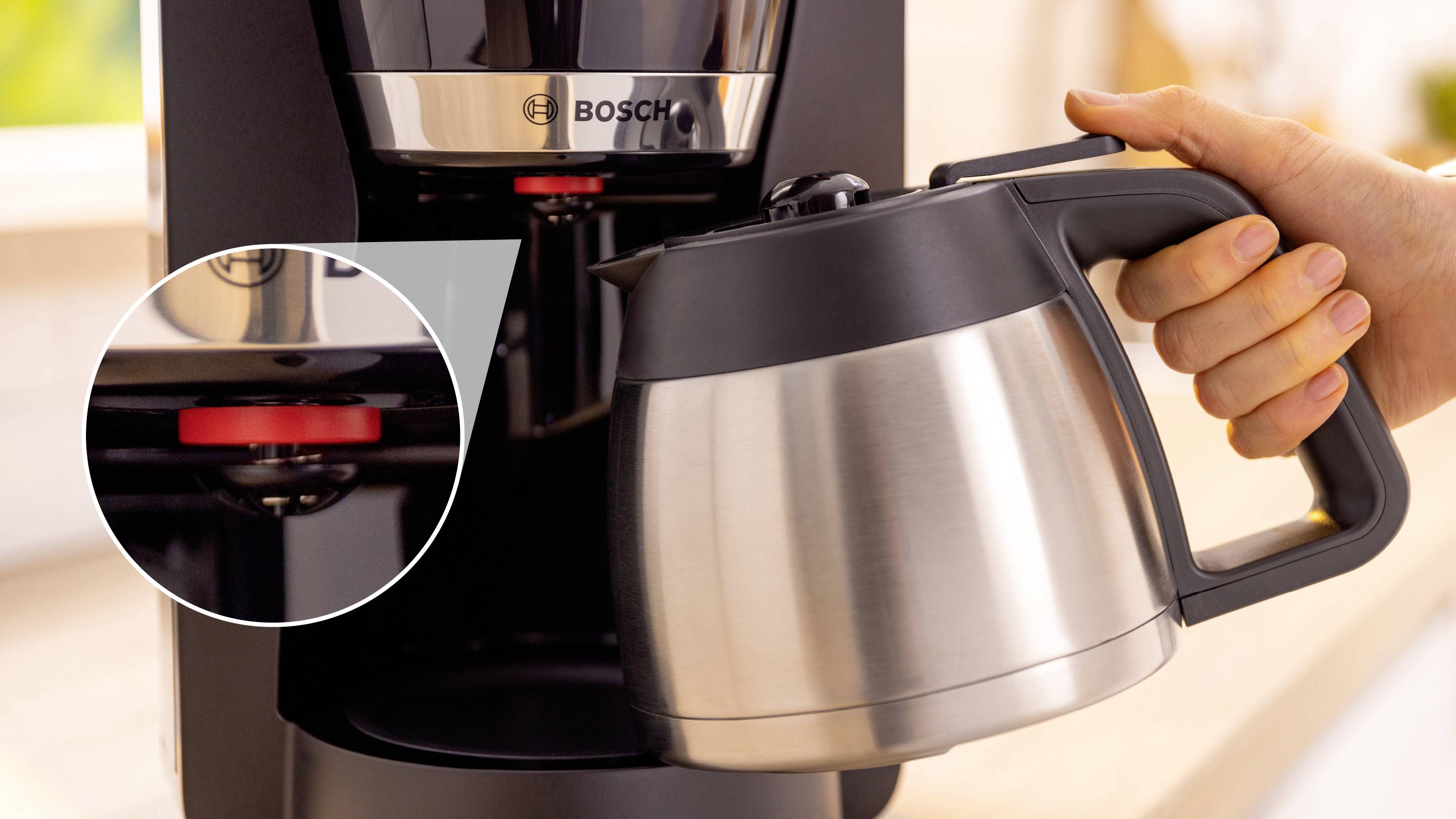 Eine Hand, die einen Kaffeekessel aus Edelstahl hält, wird unter eine Kaffeemaschine gestellt. Eine Nahaufnahme zeigt einen roten Knopf an der Maschine.