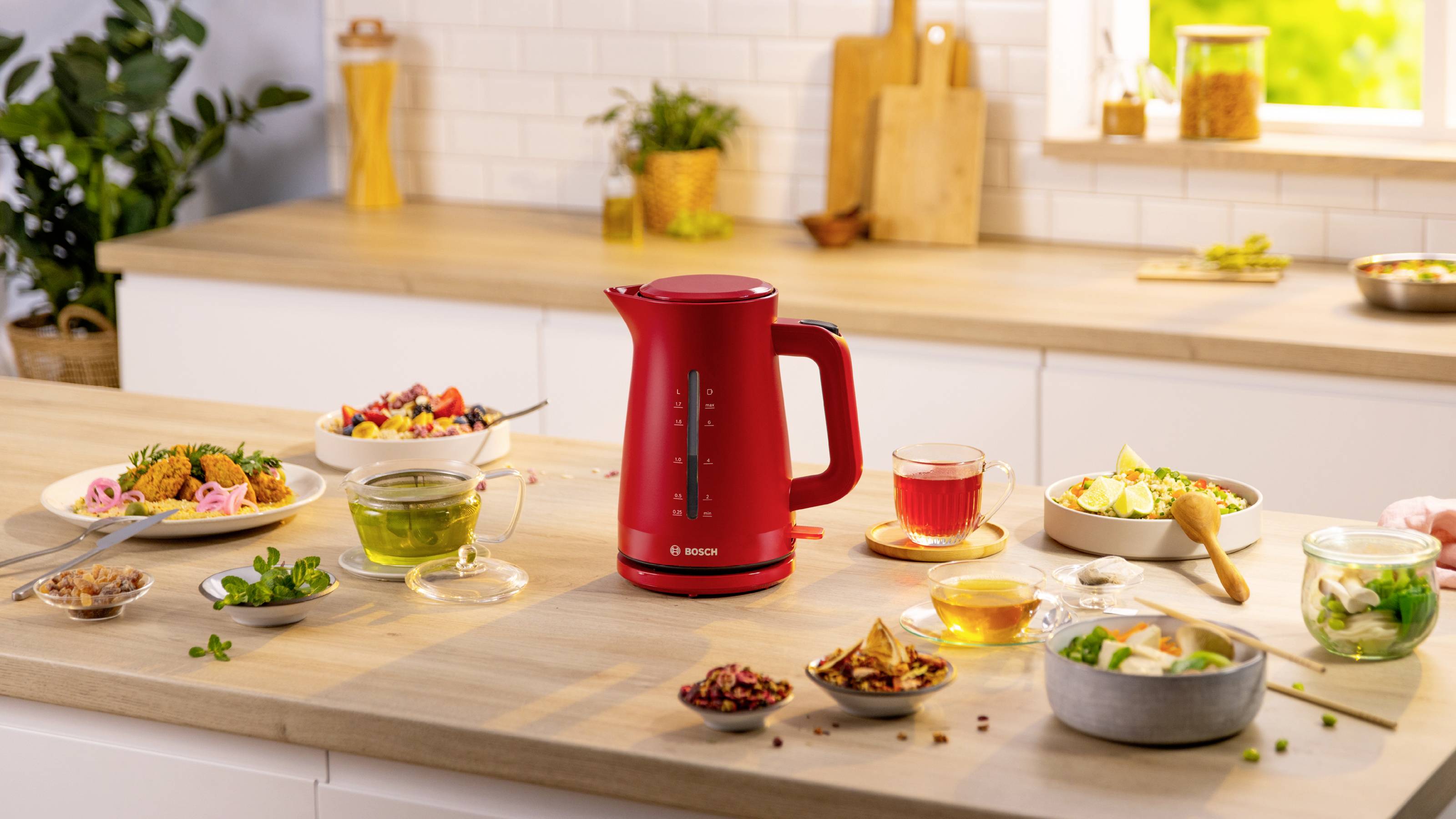 Ein roter elektrischer Wasserkocher steht auf einer Küchenarbeitsplatte, umgeben von verschiedenen Geschirren und Getränken, mit einem hellen Fenster und einer Pflanze im Hintergrund.