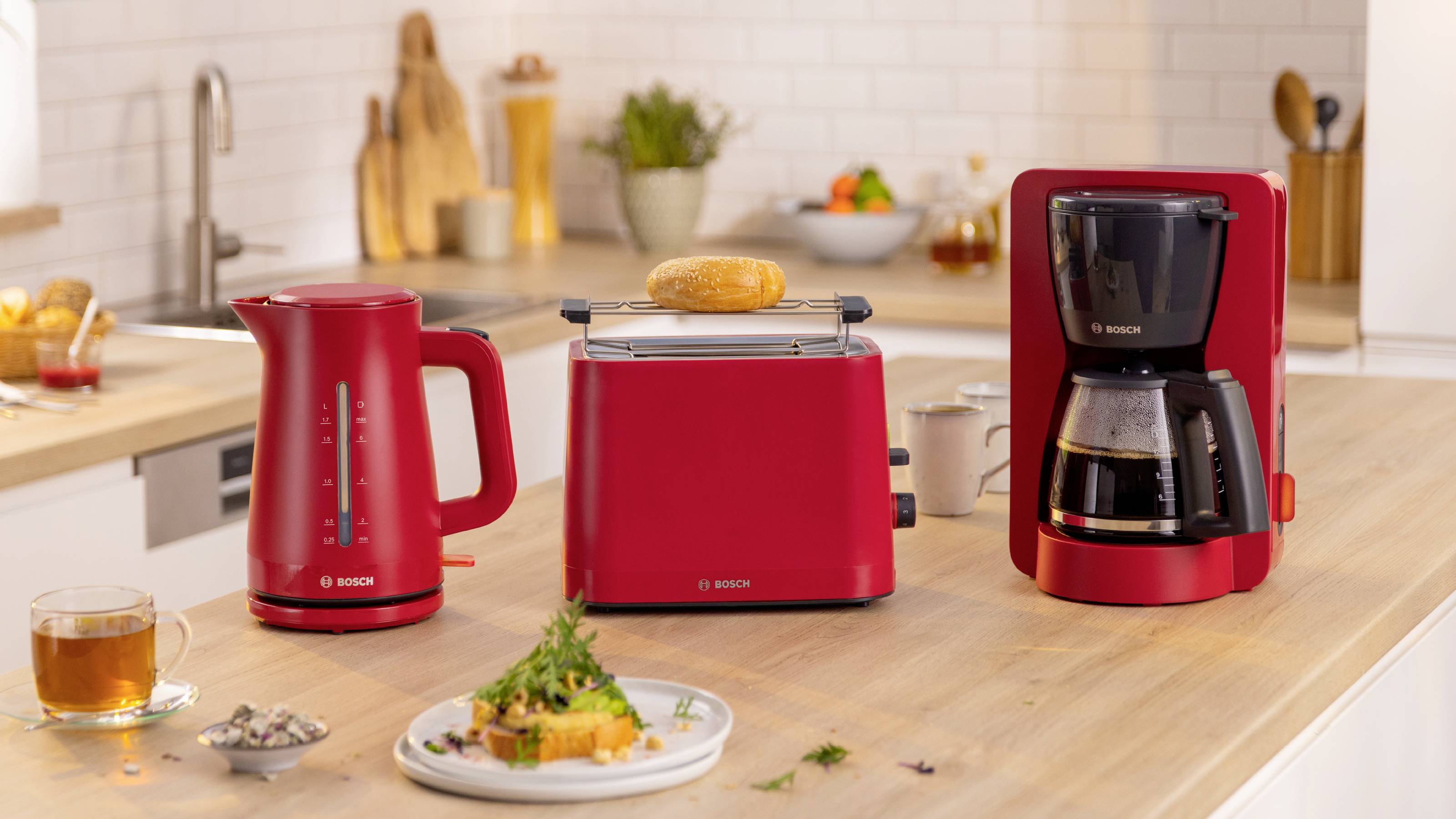 Eine Küchenarbeitsplatte mit einem roten elektrischen Wasserkocher, einem Toaster mit Toast und einer Kaffeemaschine. Daneben ein Teller mit Essen und eine Teetasse.