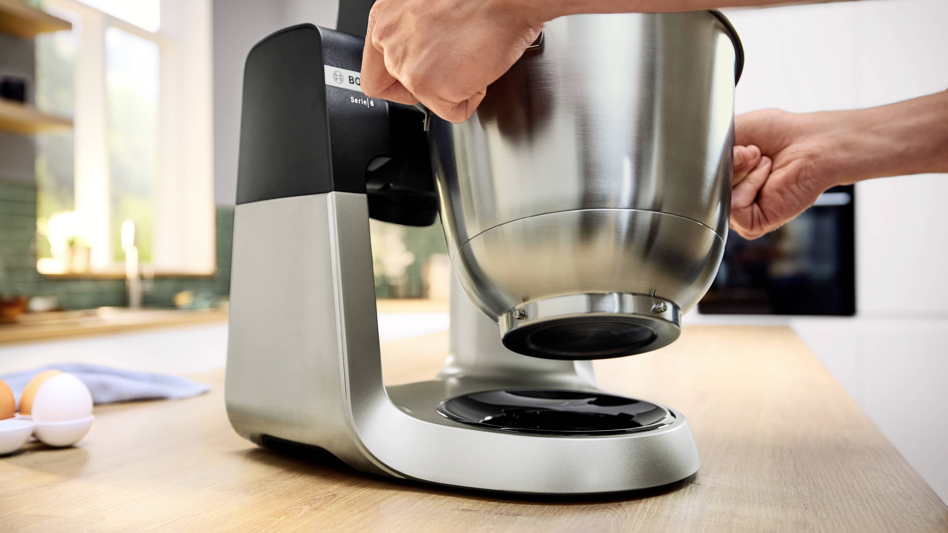 Eine Person, die eine Edelstahlrührschüssel auf einen modernen Küchenstandmixer auf einer Holzarbeitsplatte stellt. Eier und ein Handtuch befinden sich in der Nähe.