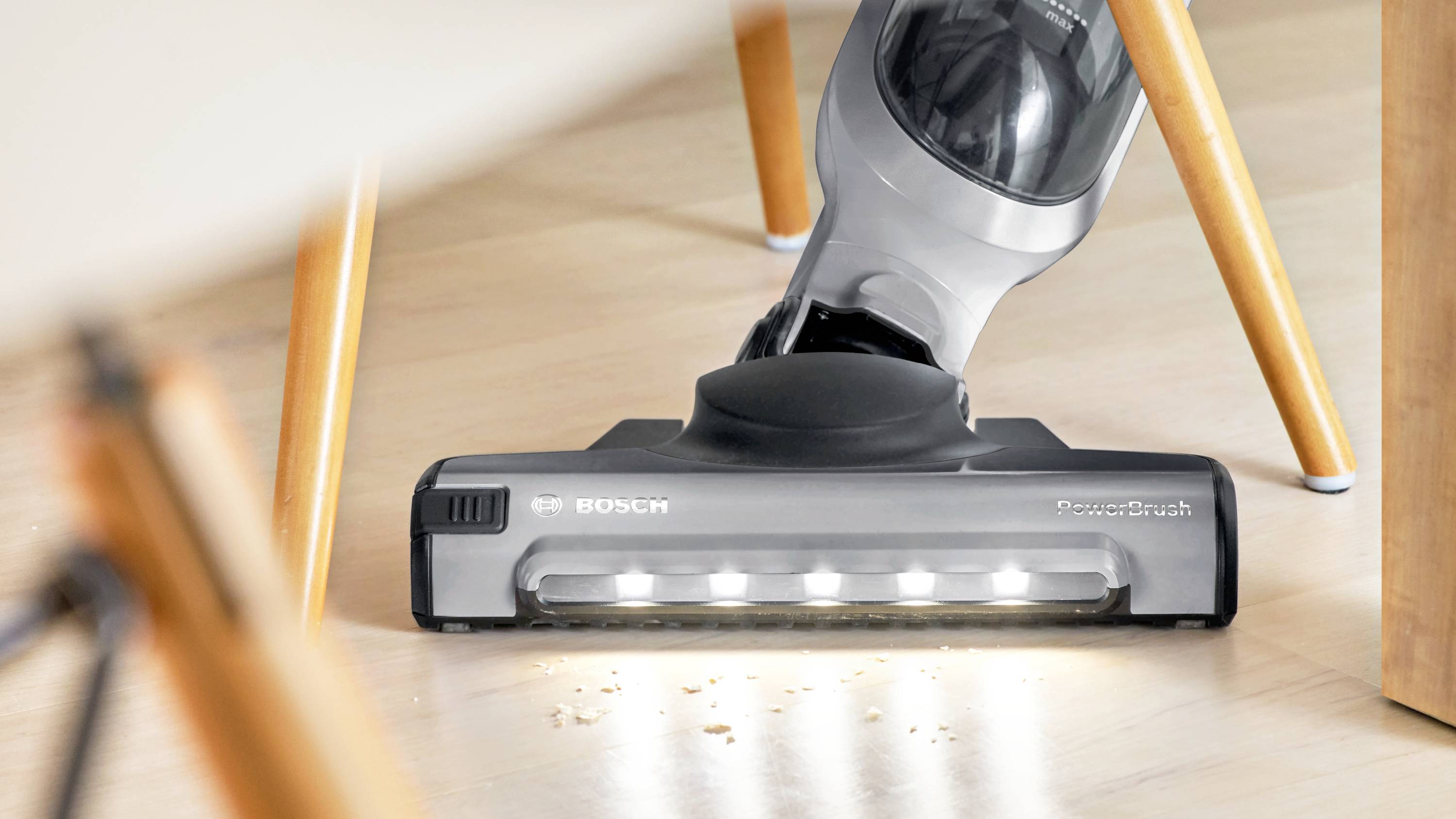 Ein Staubsauger mit LED-Beleuchtung reinigt effizient Krümel von einem glatten, hellgefärbten Boden unter einem Holzstuhl.
