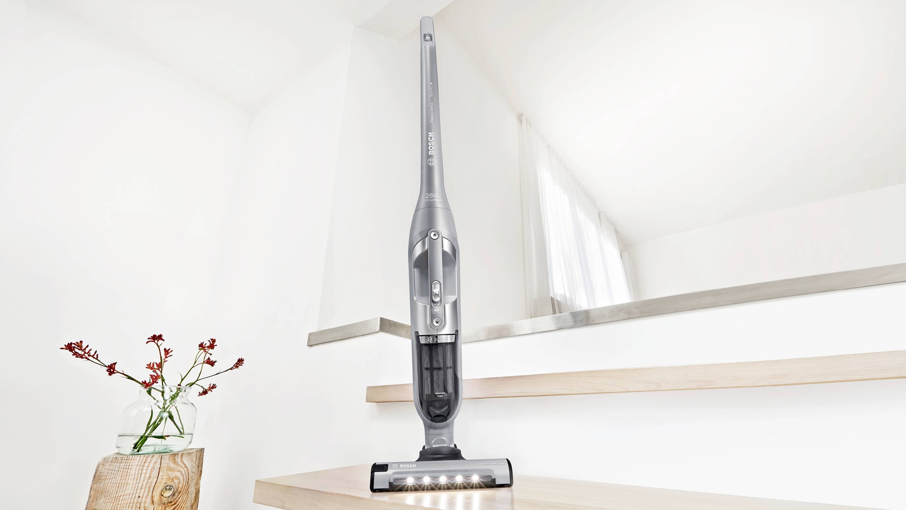 Ein kabelloses Staubsauger steht auf Holzstufen in einem hellen Raum mit einer Pflanze in der Nähe und betont dabei sein modernes, schlankes Design.