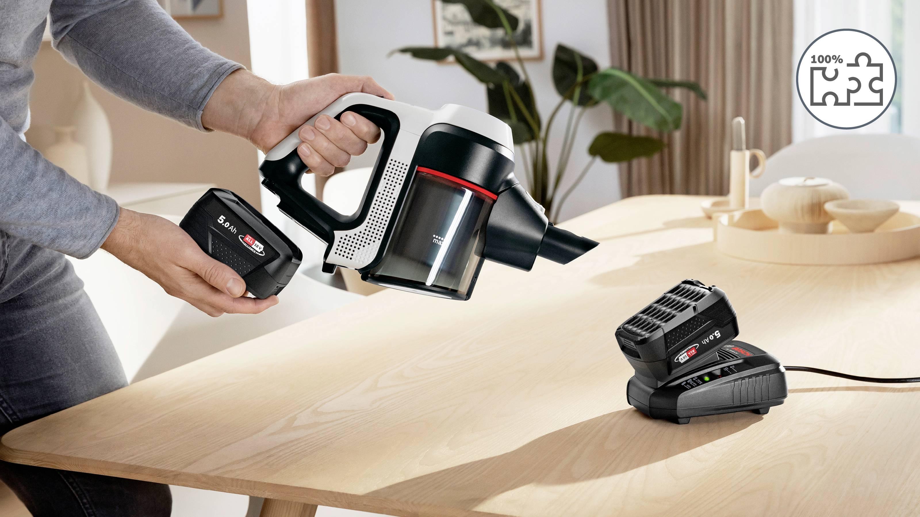 Eine Person legt eine Batterie in einen Handstaubsauger an einem Holztisch, wobei sich ein Ladegerät in der Nähe in einer Wohnumgebung befindet.