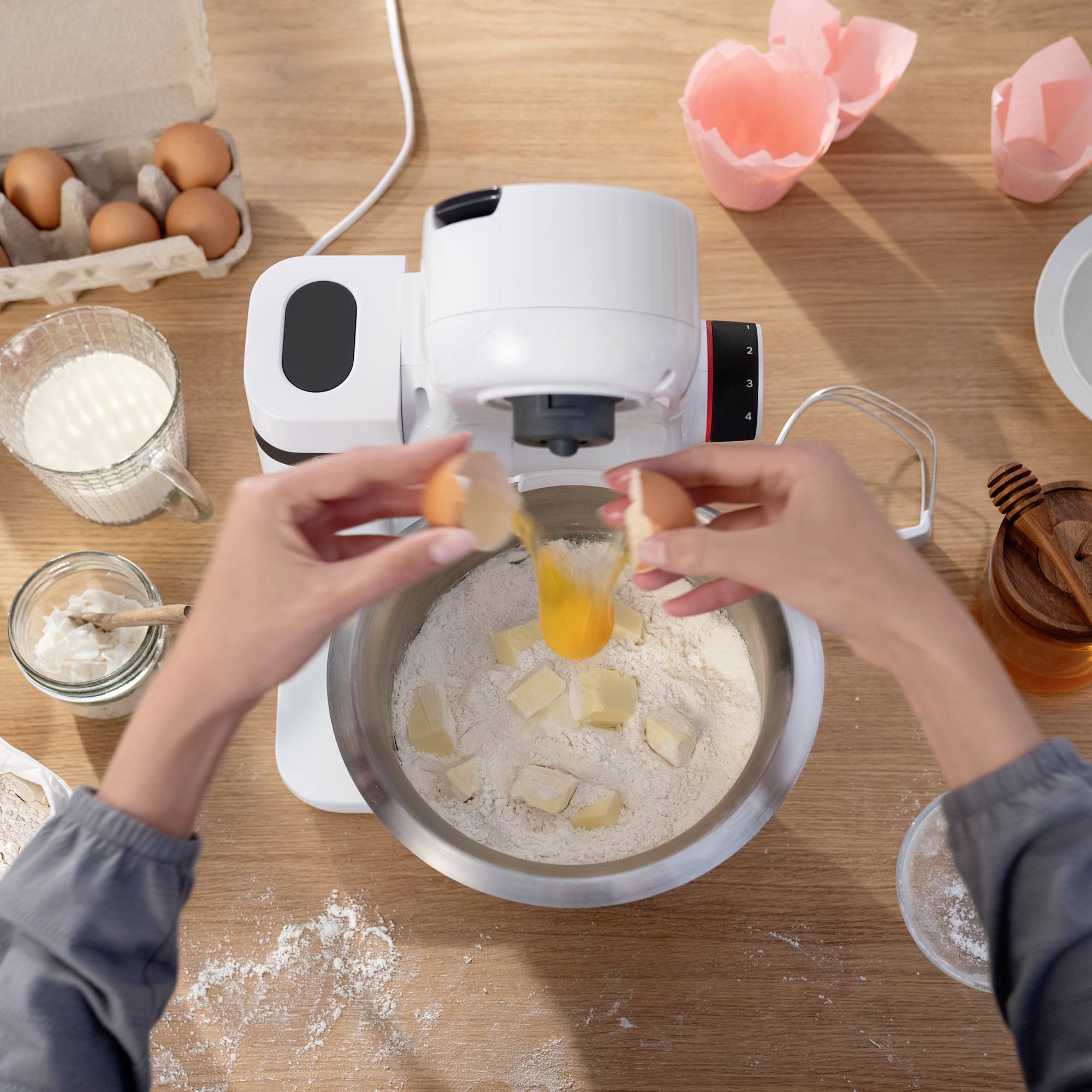 Hände, die ein Ei in eine Rührschüssel unter einem Standmixer zerbrechen, wobei sich Mehl darunter befindet. Eier, Mehl, Butter und Küchenutensilien liegen auf einer Holzoberfläche.