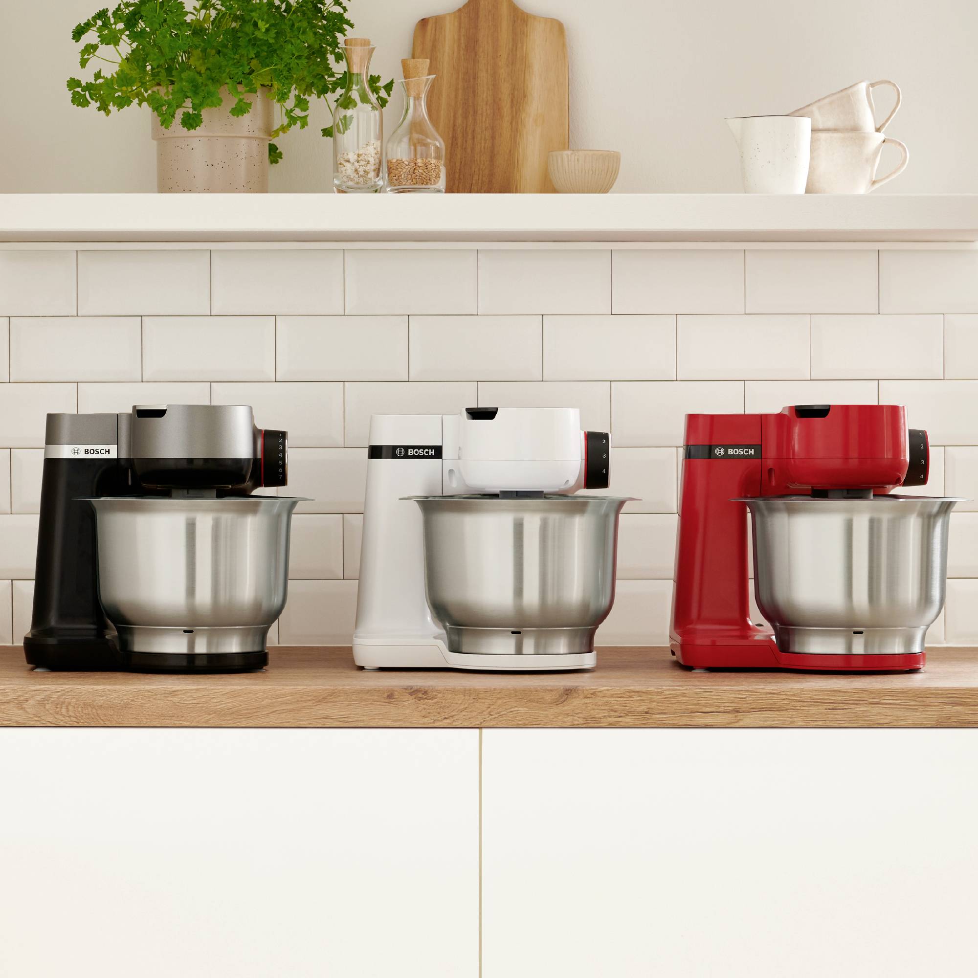 Drei Küchenmaschinen in Schwarz, Weiß und Rot auf einer Küchenarbeitsplatte unter einem Regal mit Pflanzen und Tassen vor einer weißen gefliesten Wand.