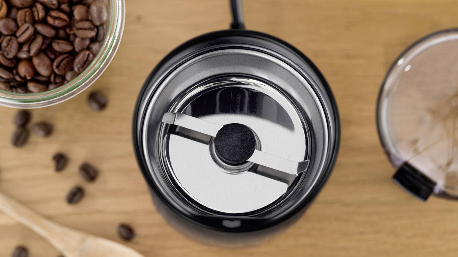 Draufsicht einer Kaffeemühle mit verstreuten Bohnen auf einer Holzoberfläche, wobei die Klinge und der Deckel der Mühle sichtbar sind.