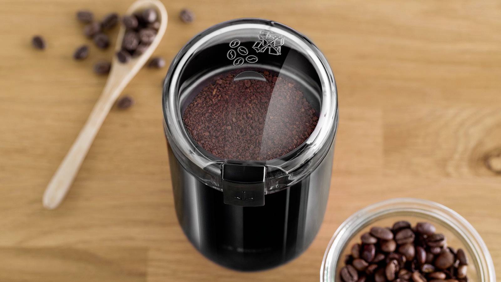 Eine schwarze Kaffeemühle, gefüllt mit gemahlenem Kaffee auf einer Holzoberfläche, umgeben von verstreuten Kaffeebohnen und einem Holzlöffel.