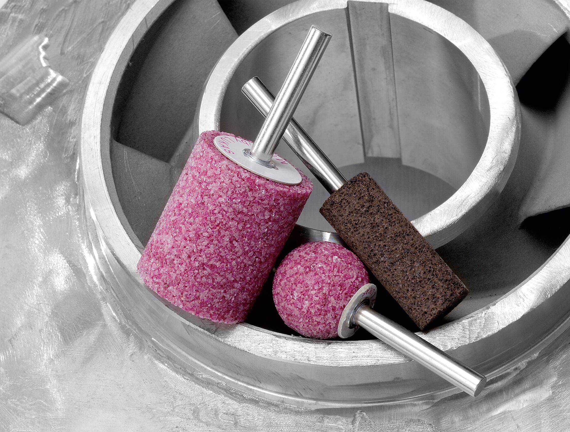 Schleifwerkzeuge verschiedener Formen auf einem metallischen, industriellen Hintergrund, mit Hervorhebung eines abrasiven rosa Rades, einer Kugel und eines braunen Blocks.