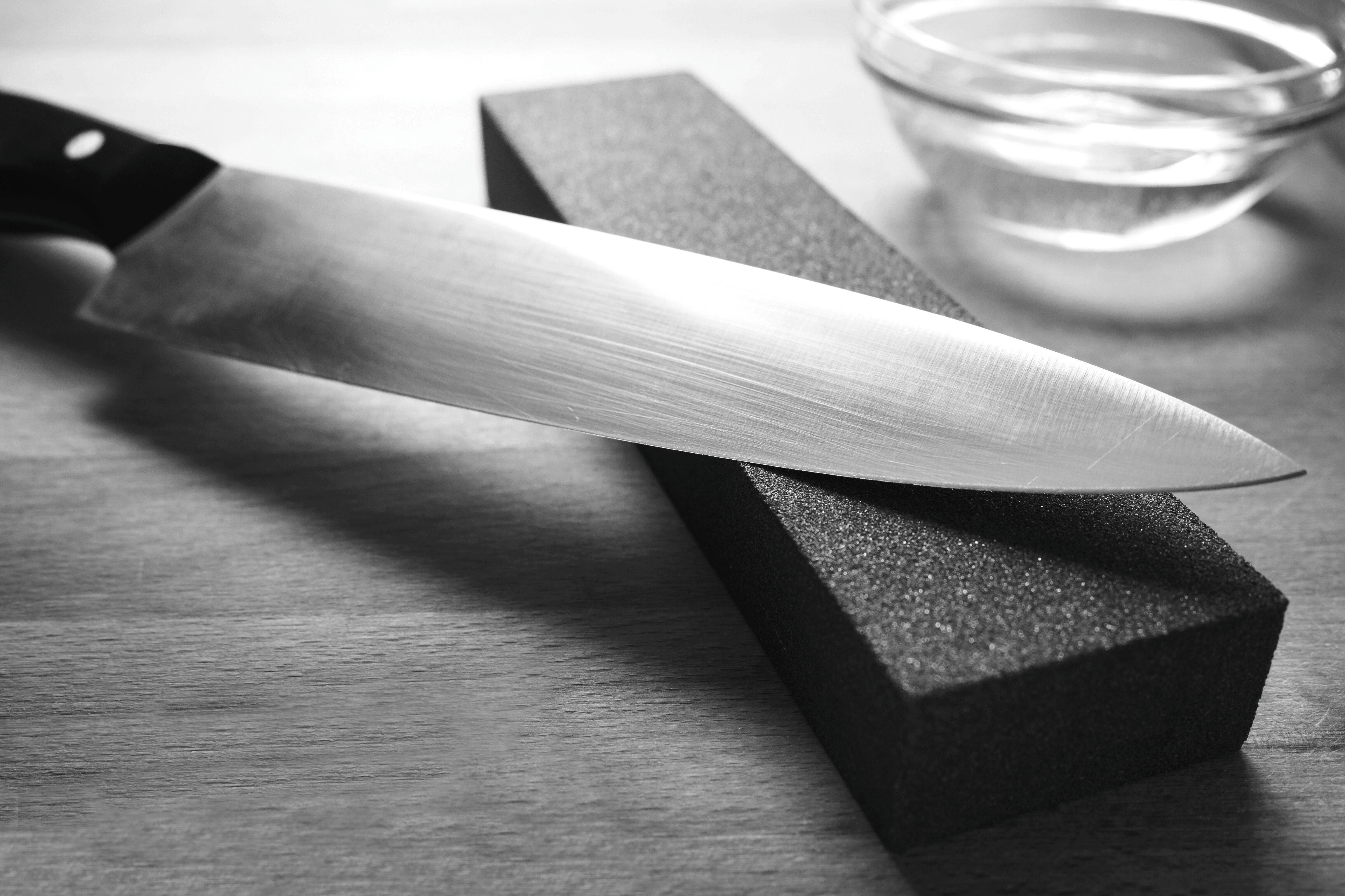 Ein scharfes Küchenmesser liegt auf einem Schleifstein, neben dem ein Glasgefäß mit Wasser auf einer Holzoberfläche steht, was auf einen Schärfvorgang hindeutet.