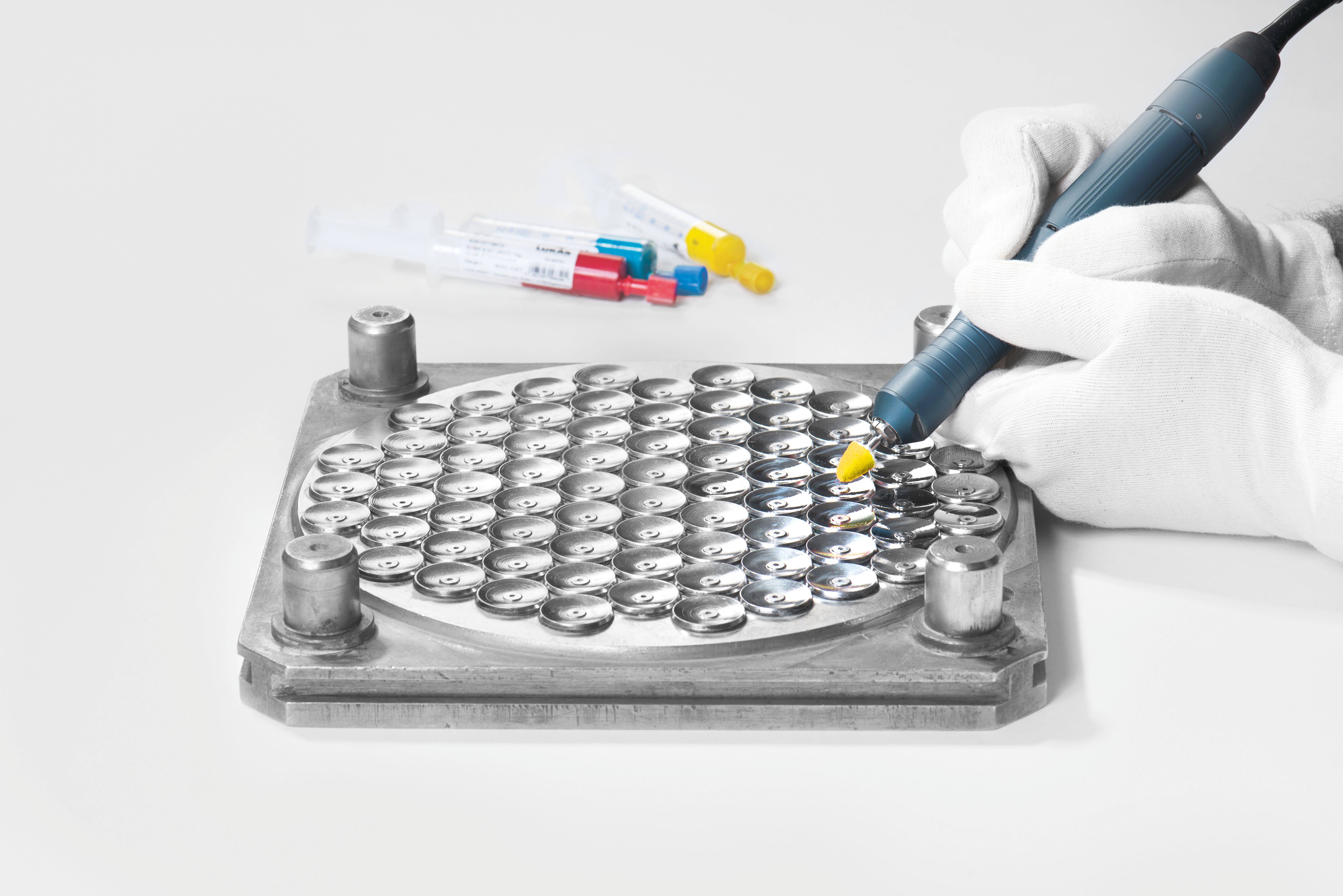 Eine Handschuh tragende Hand verwendet eine Pipette auf einem Tablett mit kleinen runden Batterien. Farbige Spritzen befinden sich im Hintergrund.