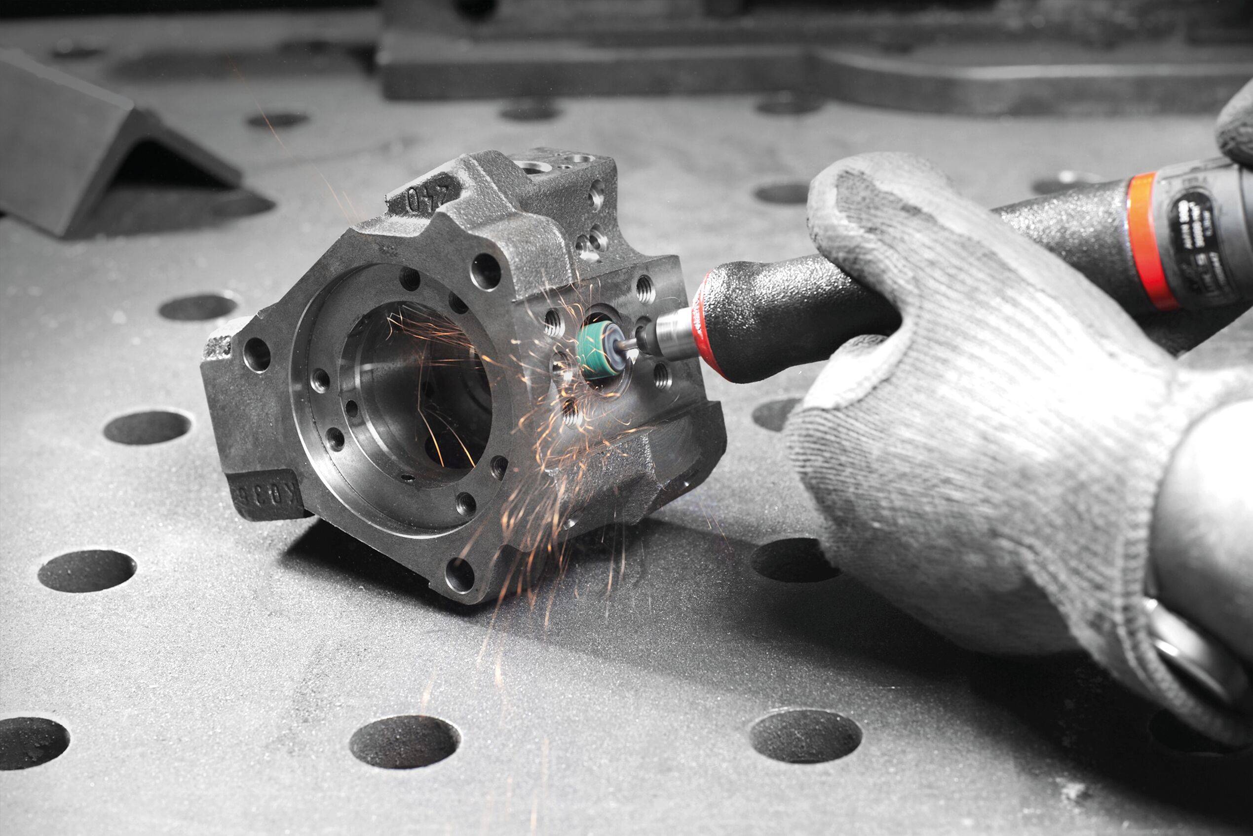 Eine Person mit Handschuhen verwendet einen Rotationsschleifer, um eine Metallkomponente zu polieren, wobei Funken durch die Luft sprühen, auf einer gelochten Metallarbeitsbank.