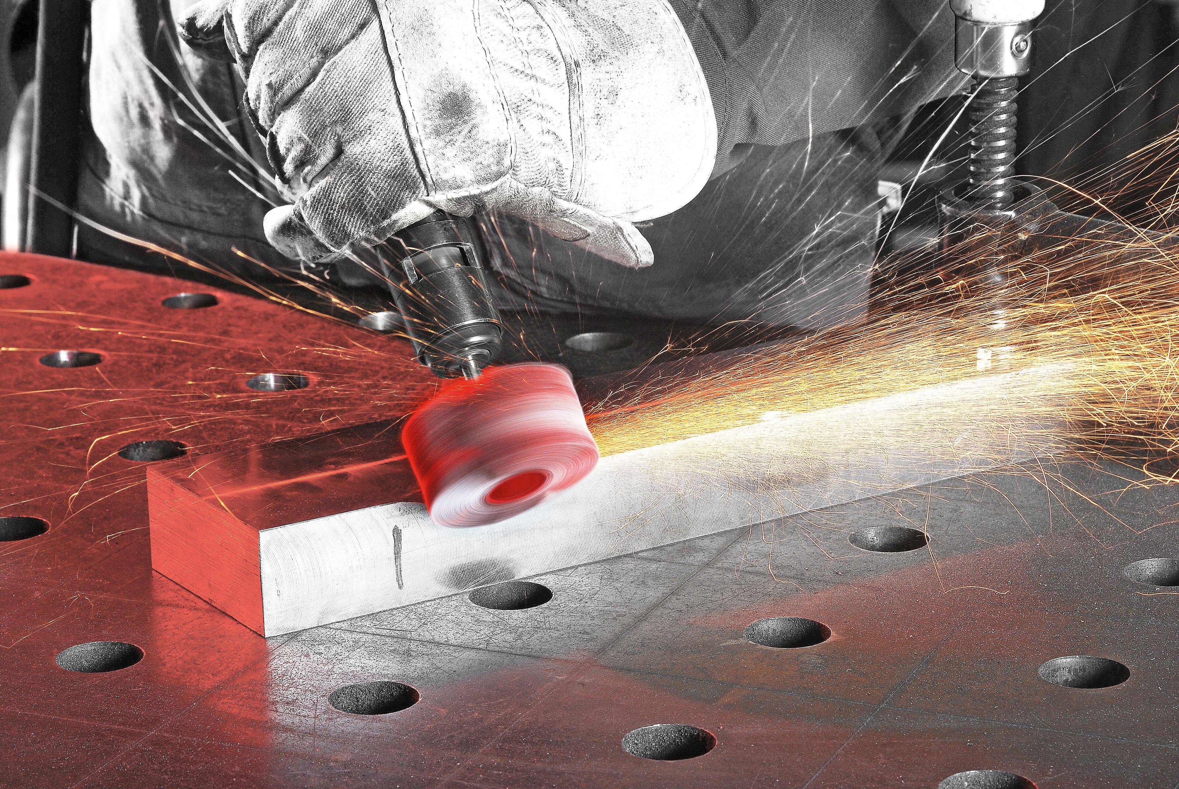 Eine Person mit Handschuhen verwendet ein Rotationswerkzeug, um einen Metallblock auf einer gelochten Werkbank zu schleifen und dabei helle Funken zu erzeugen.