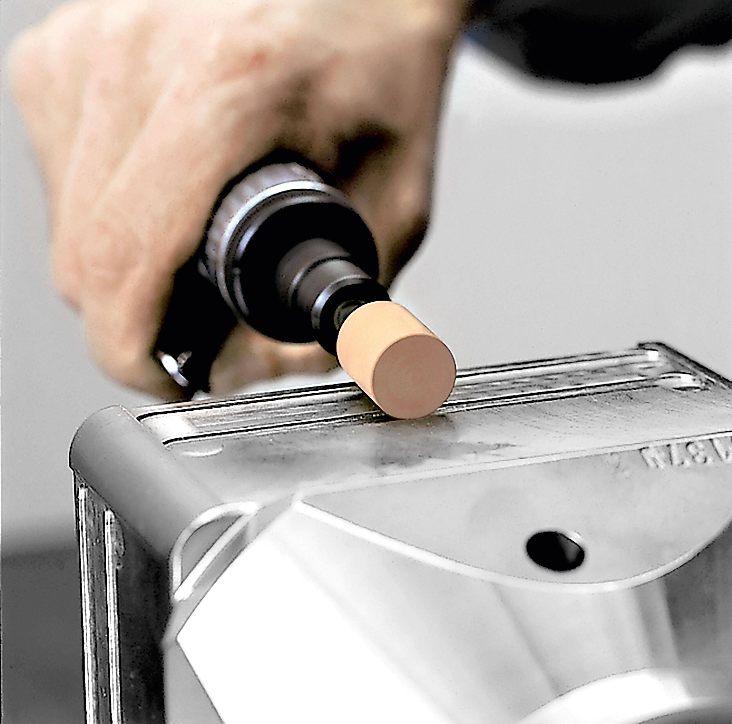 Eine Hand hält ein zylindrisches Werkzeug und drückt einen Korken in den Hals eines Metallobjekts, vermutlich eine Flasche oder einen Behälter, auf einer Werkbank.