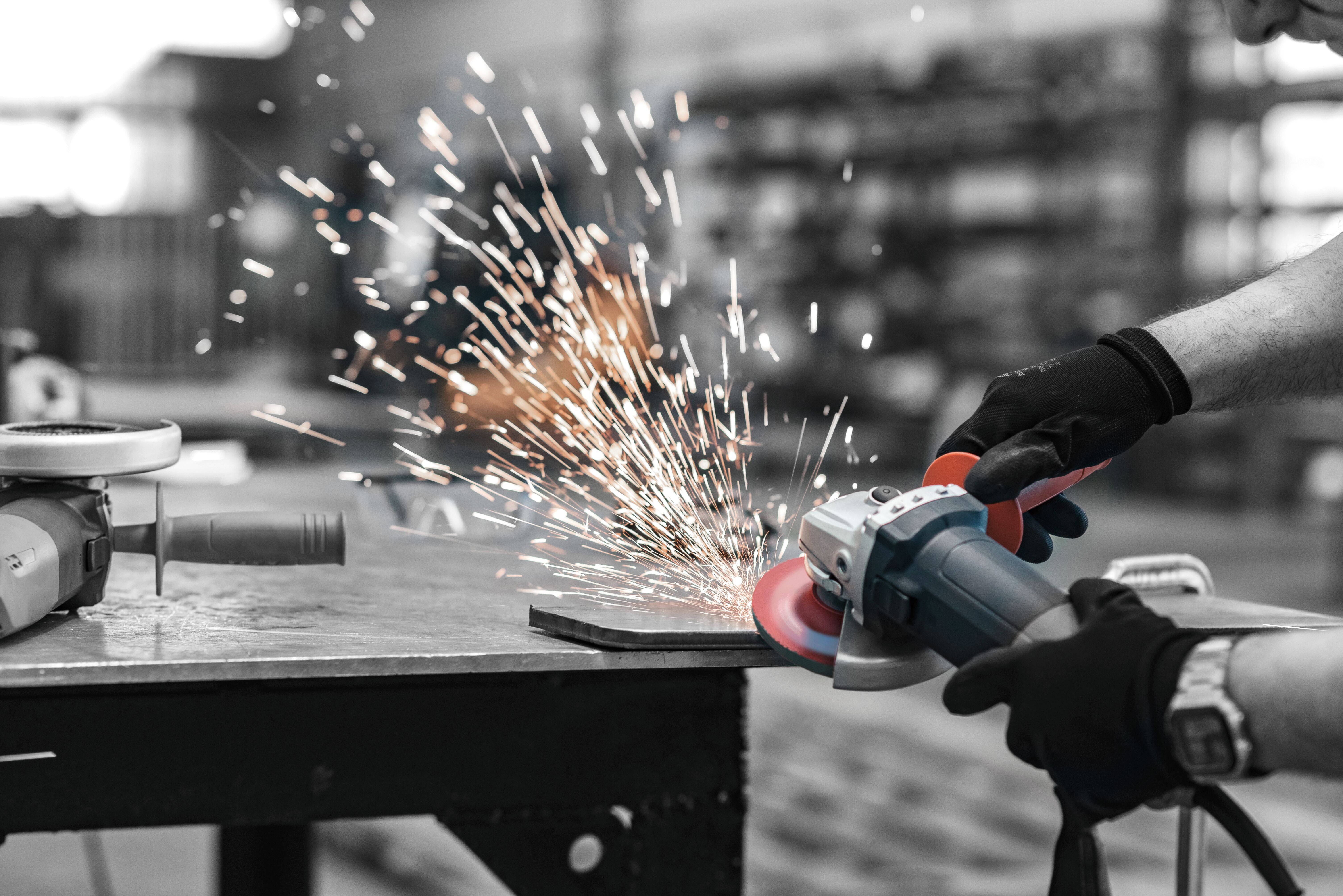 Eine Person mit Handschuhen bedient einen Winkelschleifer und erzeugt dabei helle Funken an einem Metallstück in einer Werkstattumgebung.