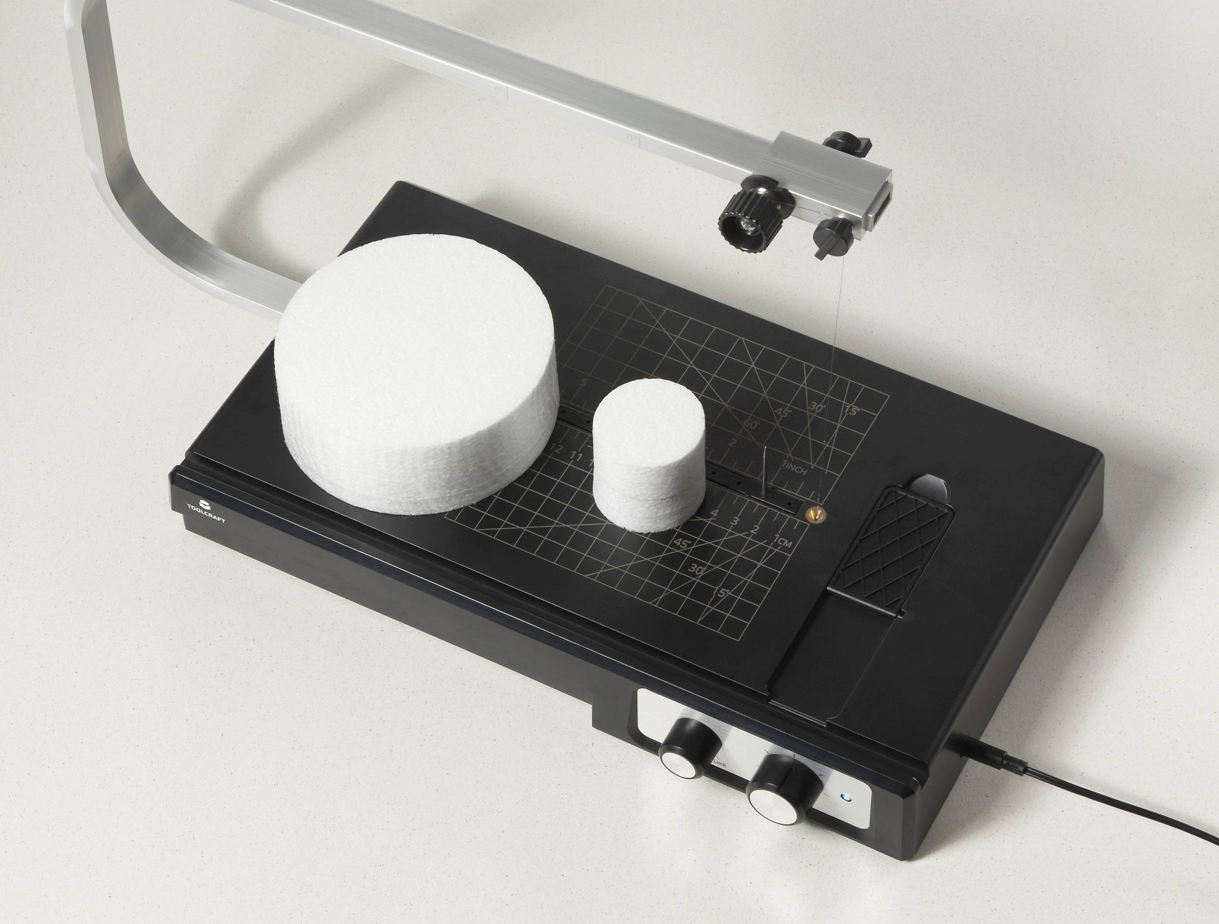 Ein Heißdraht-Schaumschneider auf einem Tisch, dargestellt mit zwei Schaumstoffstücken – einem großen und einem kleinen. Das Gerät verfügt über eine Gitterarbeitsfläche.