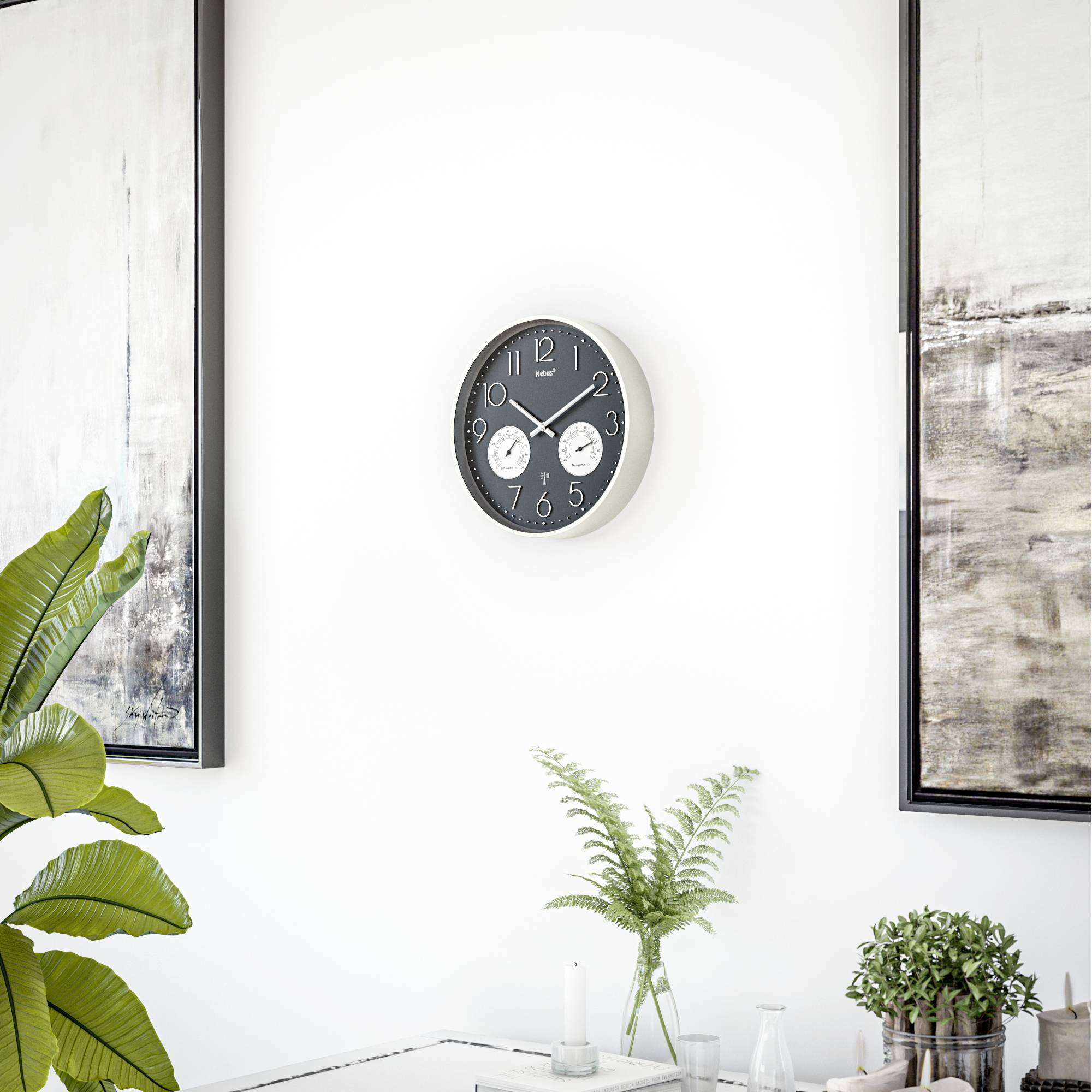 Ein minimalistisches Zimmer zeigt eine Wanduhr mit zwei kleineren Zifferblättern, gerahmte abstrakte Kunst und einen Tisch mit Farnen und Kerzen.