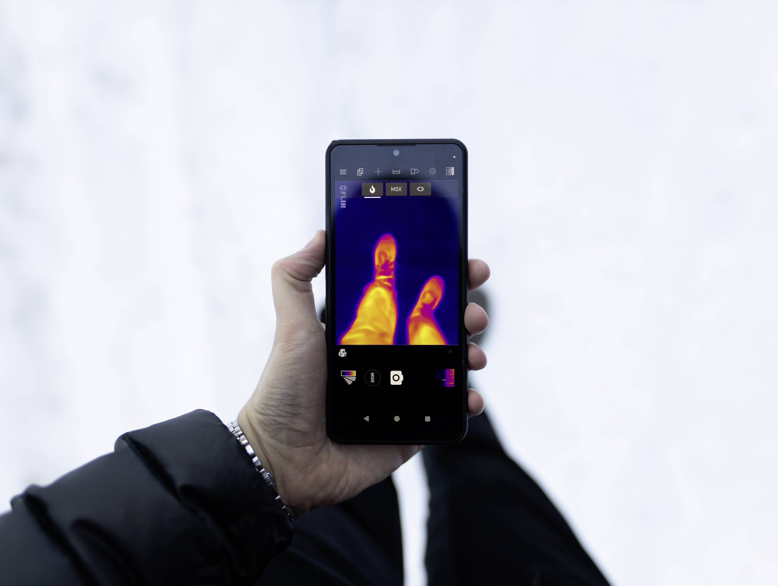 Eine Person hält ein Smartphone, das ein Wärmebild ihrer Füße zeigt, wobei der Wärmekontrast vor einem verschneiten Hintergrund hervorgehoben wird.