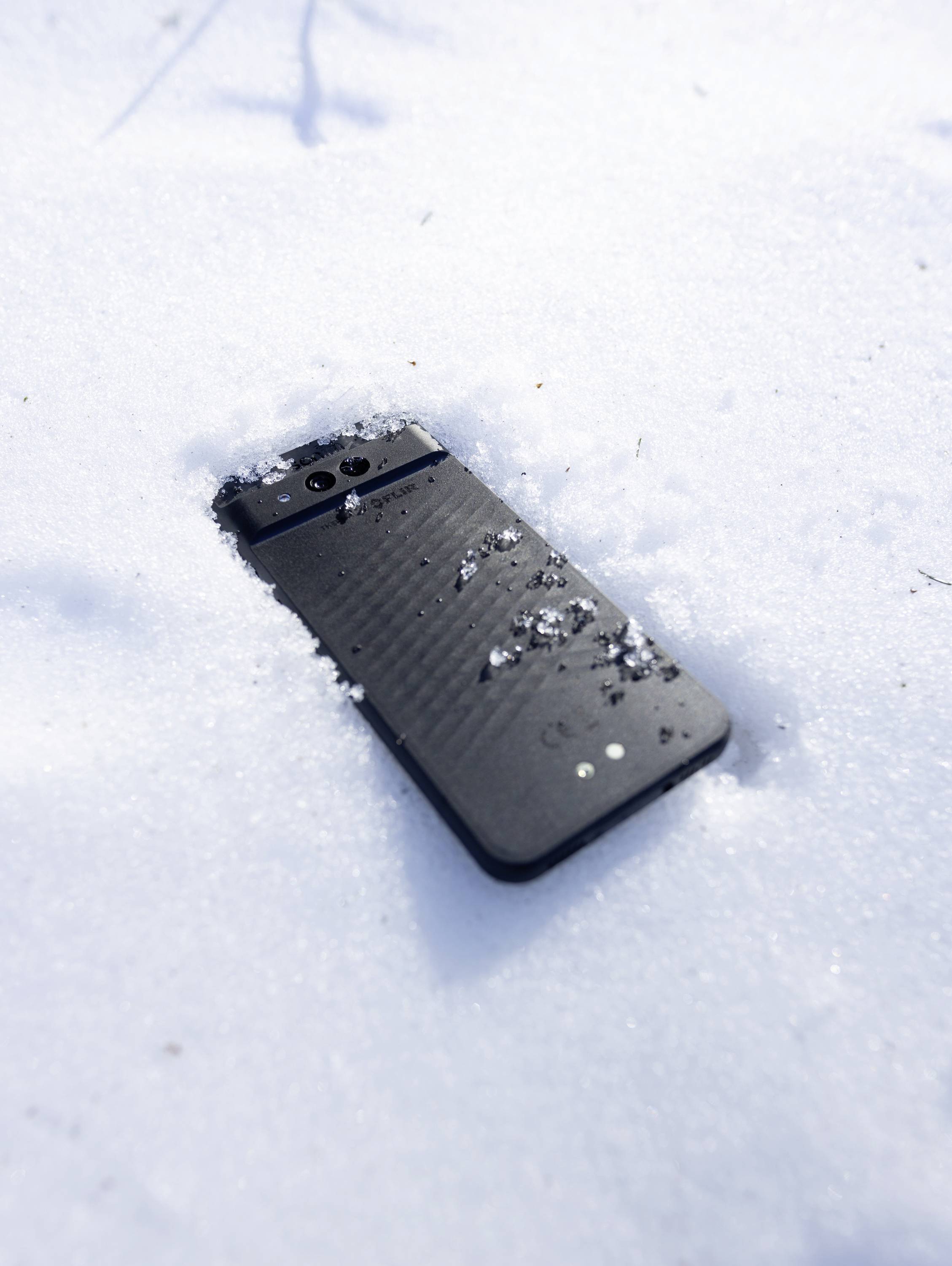 Ein Smartphone liegt teilweise im Schnee vergraben, mit einigen Schneeflocken auf seiner Oberfläche, was auf kalte Wetterbedingungen hindeutet.