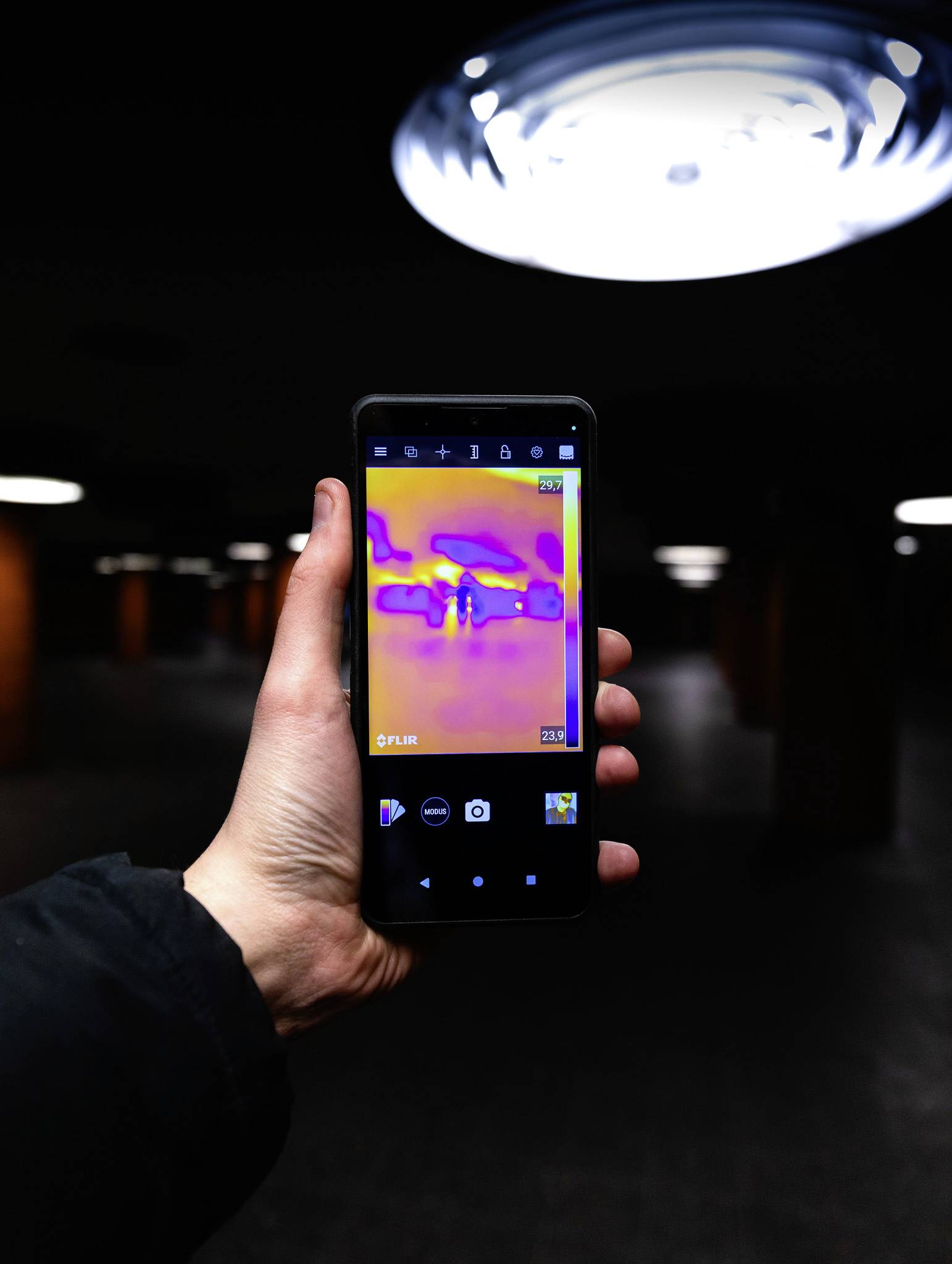 Eine Hand hält ein Telefon, das in einer dunklen Umgebung ein Wärmebild zeigt, während helle Deckenlichter im Hintergrund zu sehen sind.