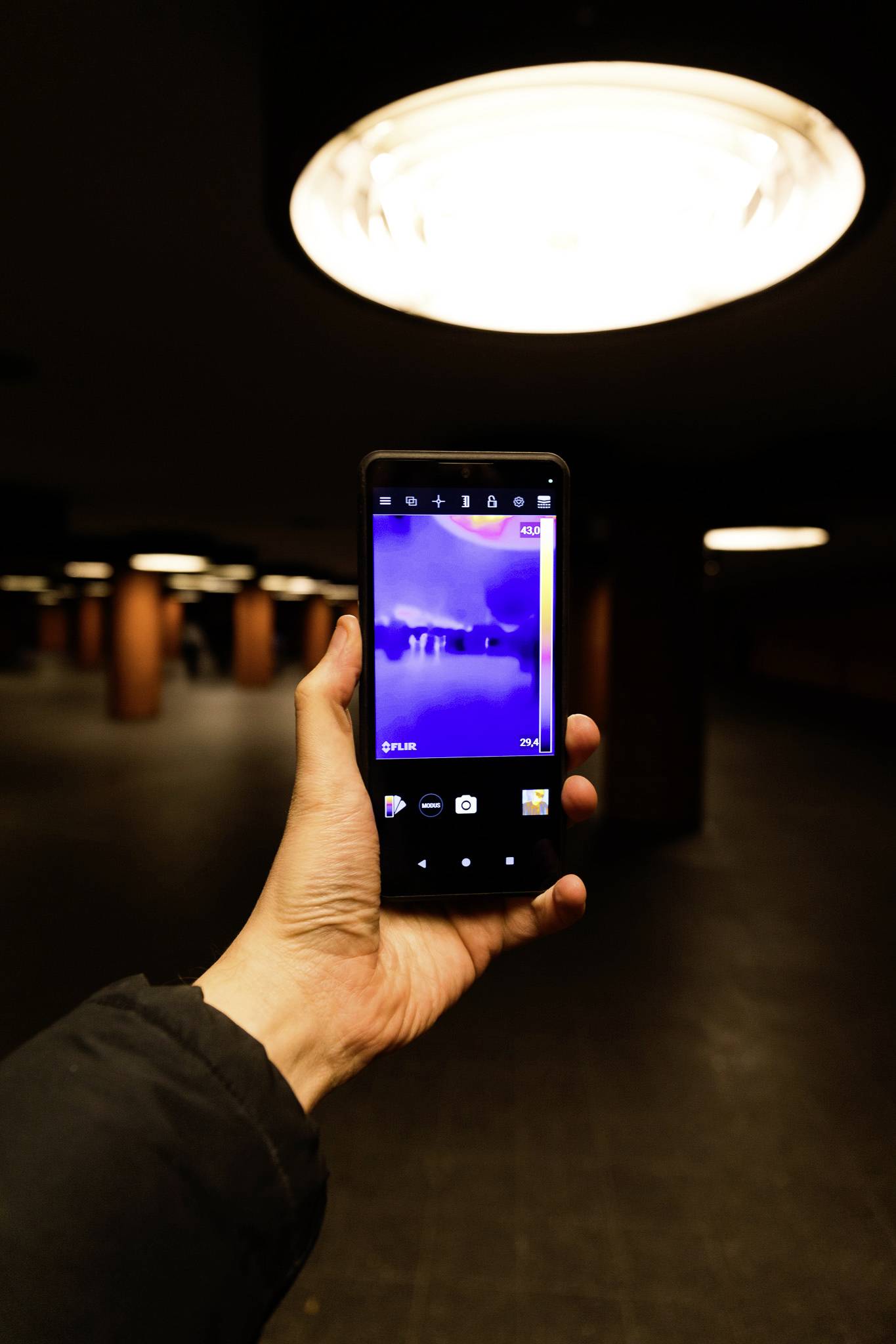 Eine Person hält ein Smartphone, auf dem eine Wärmebildkamera-Ansicht in einem dunkel beleuchteten Innenraum mit runden Deckenleuchten und Säulen zu sehen ist.