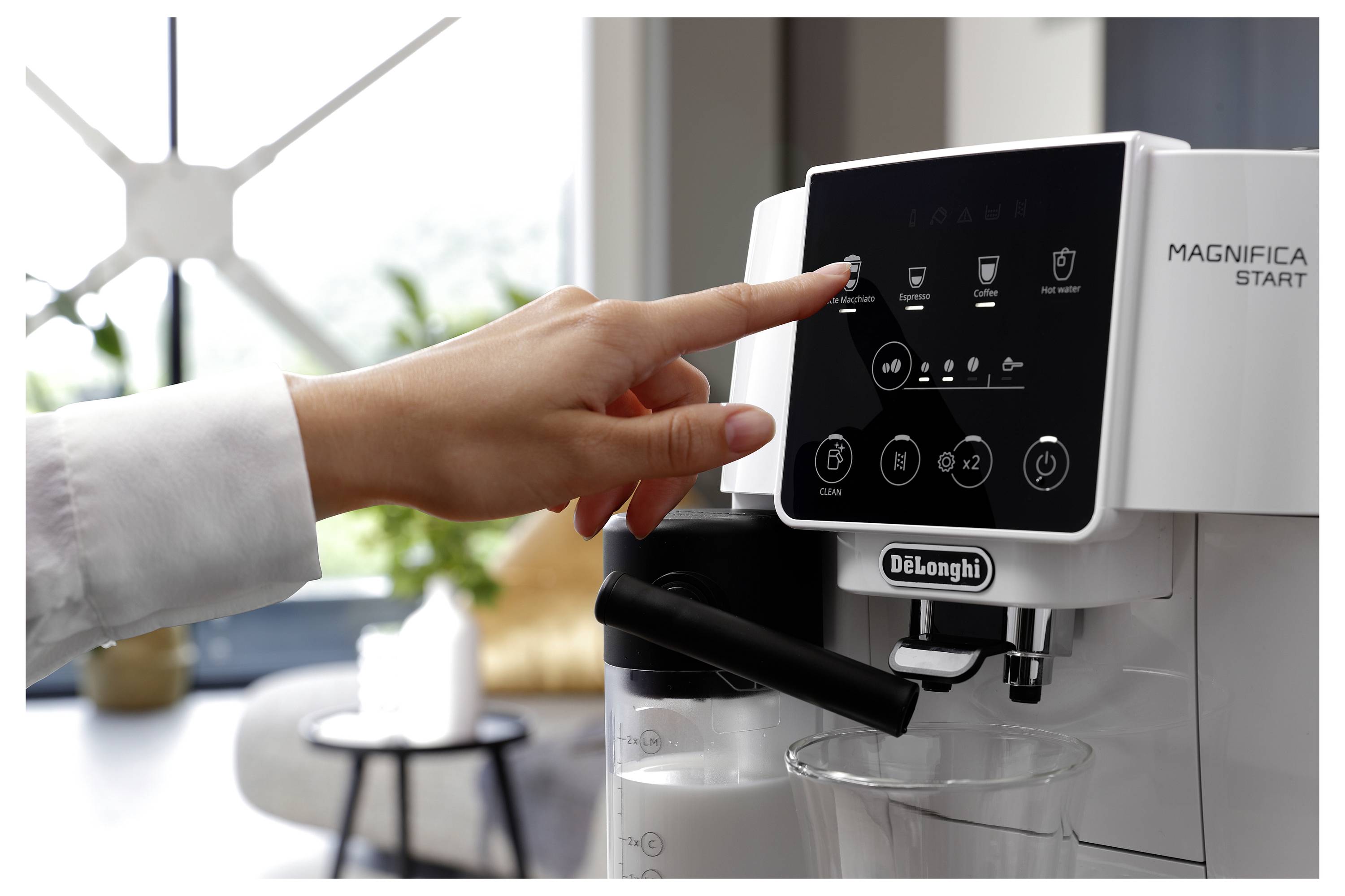 Eine Hand wählt auf dem Touchscreen einer DeLonghi Magnifica Start Espressomaschine eine Kaffee-Option aus, mit einem modernen Wohnzimmer im Hintergrund.