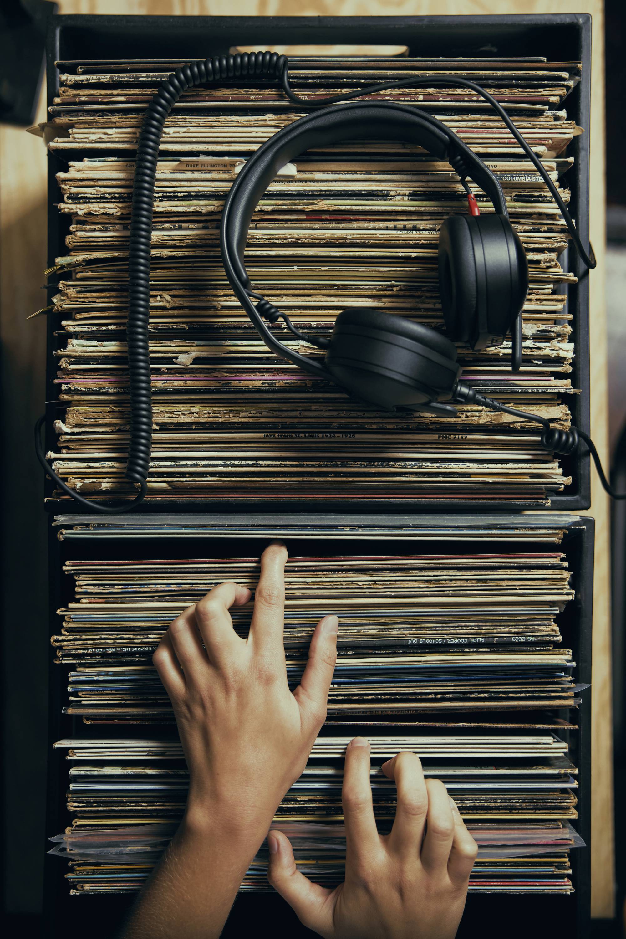 Eine Paar Hände, die durch eine Sammlung von Schallplatten in einer Kiste sortieren, mit einem Kopfhörer, der darauf ruht.