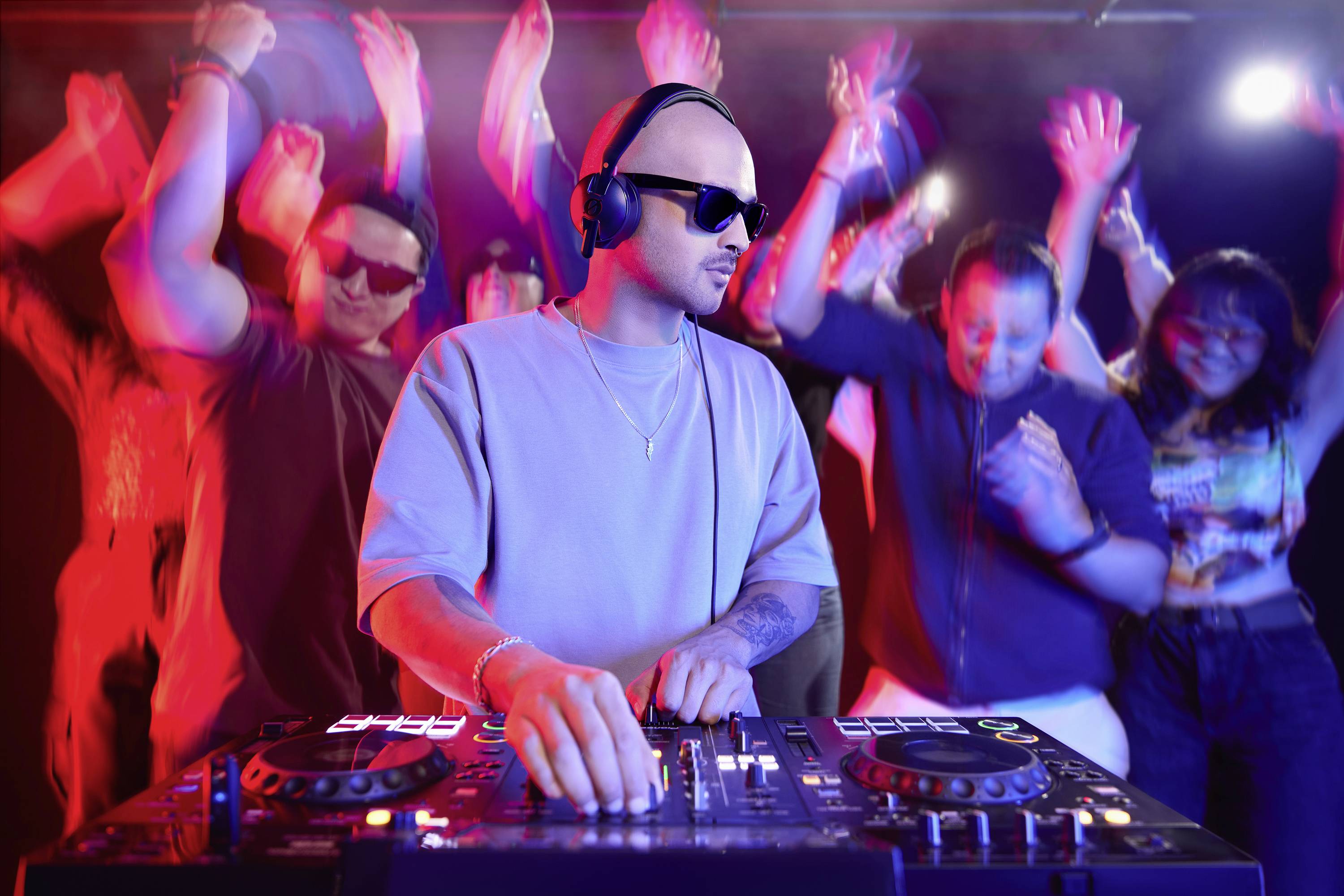 Ein DJ mit Kopfhörern mischt Musik an einem Mischpult in einem belebten Club. Menschen mit erhobenen Händen tanzen energisch in bunten Lichtern.
