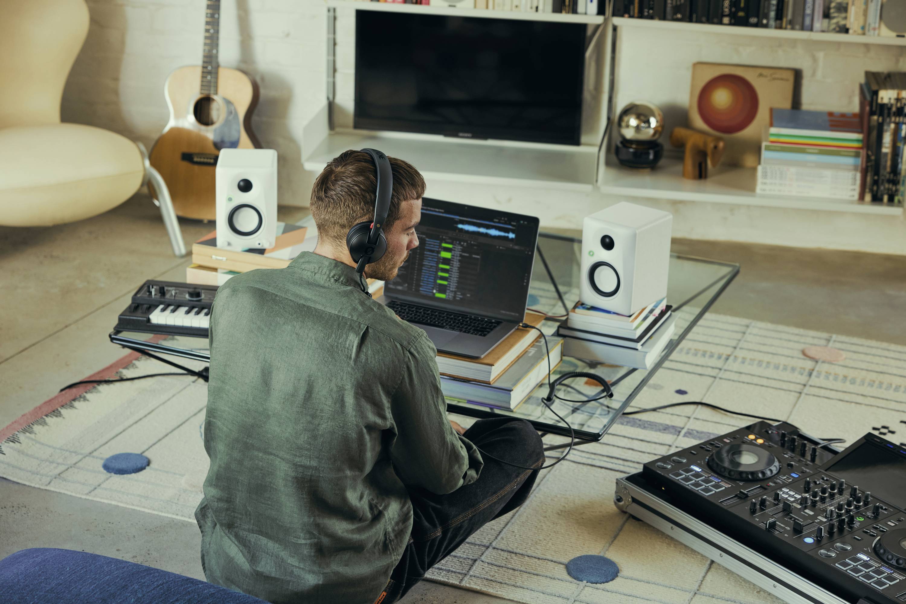 Eine Person mit Kopfhörern sitzt auf dem Boden und arbeitet an einem Laptop mit Musikproduktions-Software. Musikalische Ausrüstung umgibt den Arbeitsplatz.