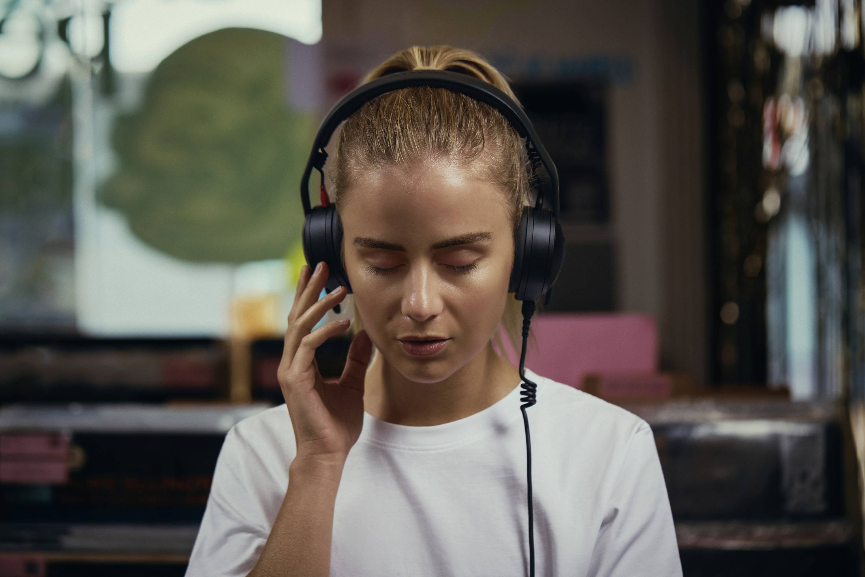 Eine Person mit geschlossenen Augen hört mit Kopfhörern Musik, steht in einem Innenraum mit unscharfem Hintergrund und vermittelt dabei ein Gefühl der Entspannung.
