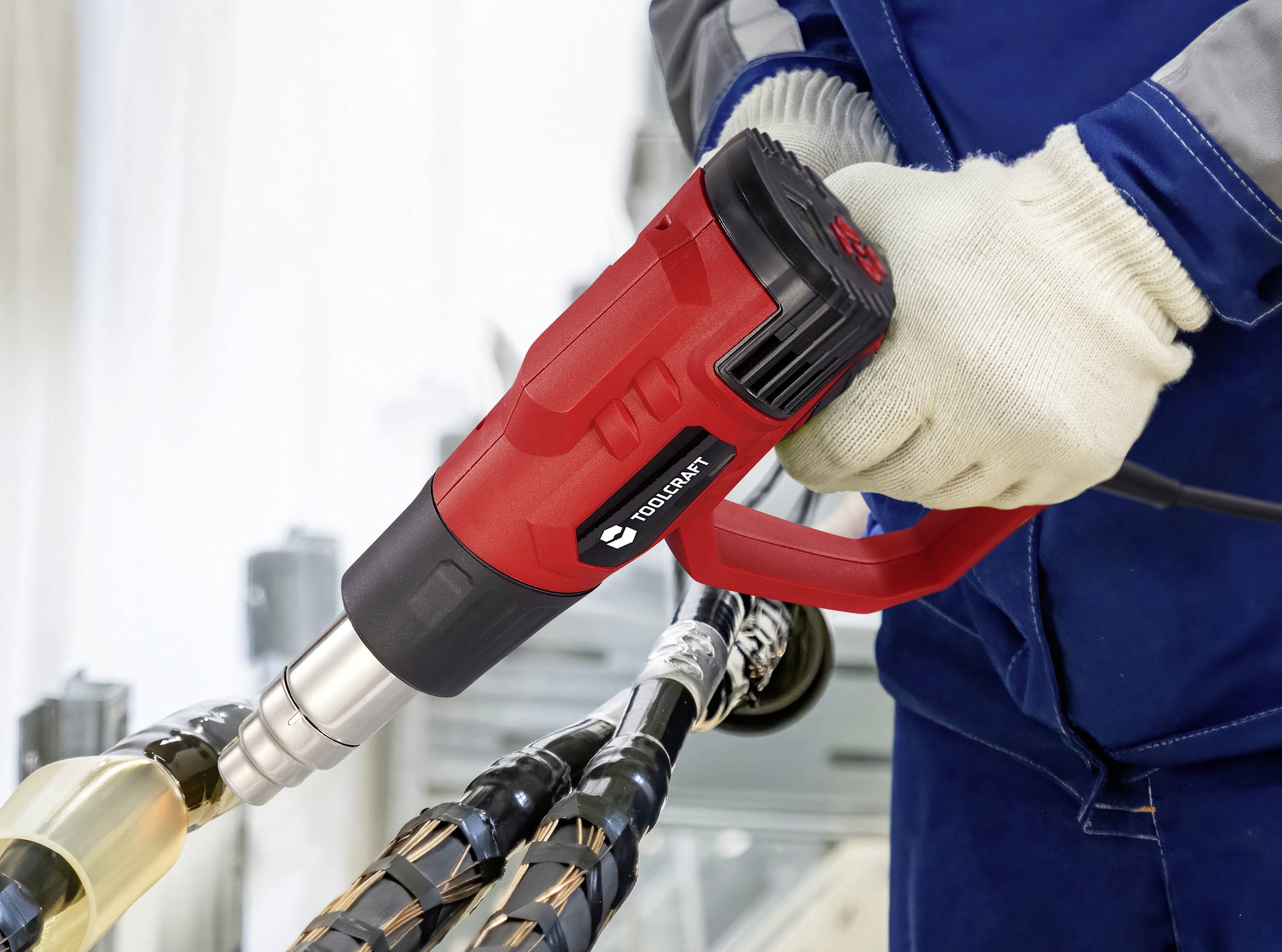 Eine Person mit Schutzhandschuhen bedient eine rote Heißluftpistole an elektrischen Kabeln, was auf eine Tätigkeit im Zusammenhang mit Kabelreparatur oder -installation hindeutet.