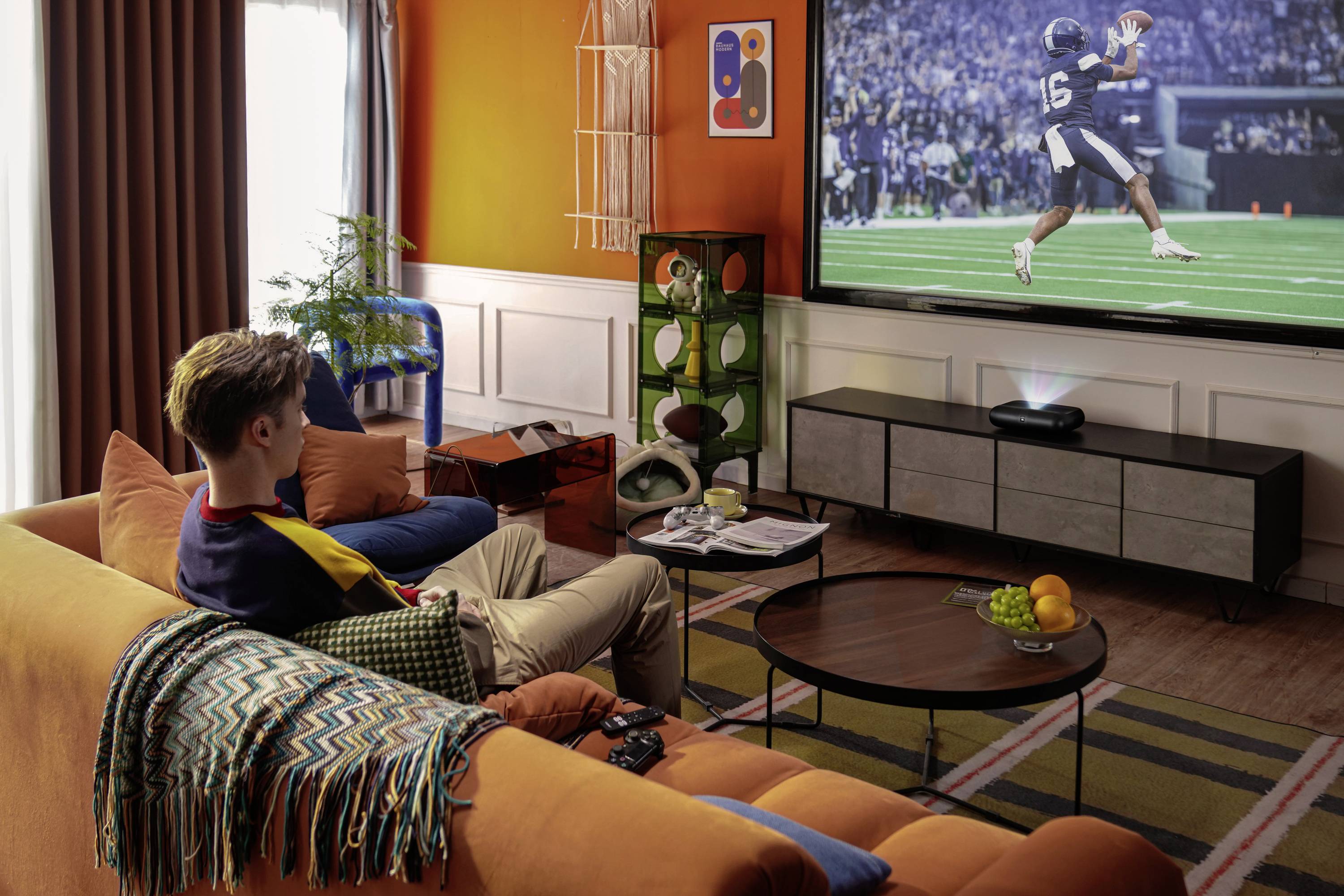 Eine Person sitzt auf einem orangefarbenen Sofa und schaut ein Fußballspiel auf einem großen Fernseher in einem gemütlichen Wohnzimmer mit lebhafter Dekoration.