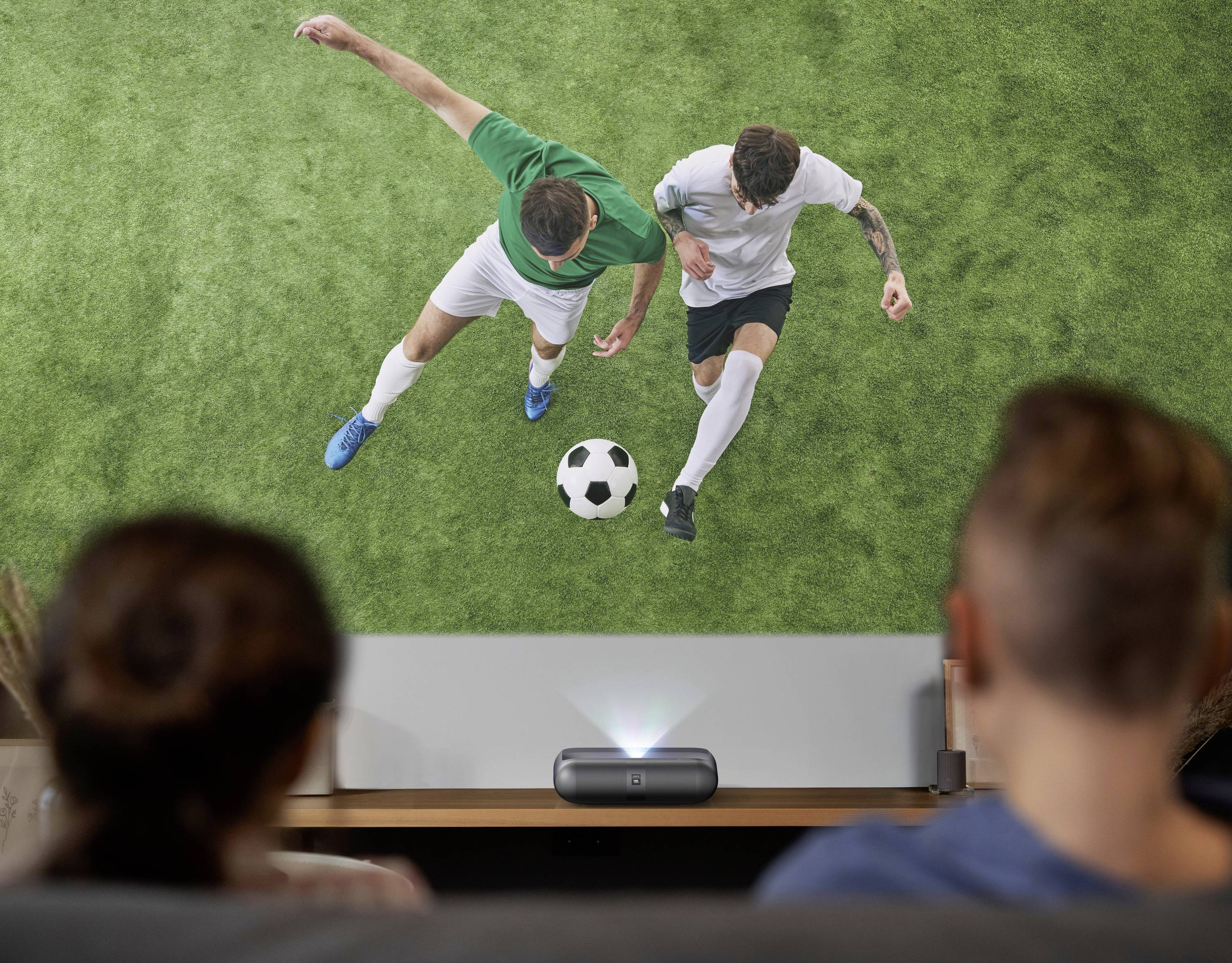 Zwei Personen beobachten ein projiziertes Bild von zwei Fußballspielern, die auf einem grasbewachsenen Feld um einen Ball kämpfen, wobei eine lebhafte Sportszene hervorgehoben wird.