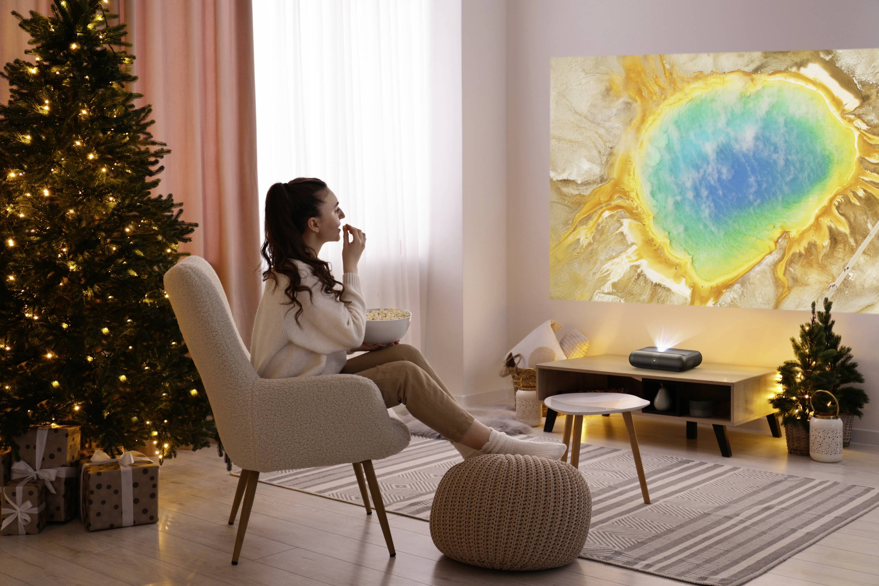 Eine Frau sitzt in einem gemütlichen Raum und beobachtet eine lebendige Naturszene, die an der Wand projiziert wird, während daneben ein geschmückter Weihnachtsbaum steht.