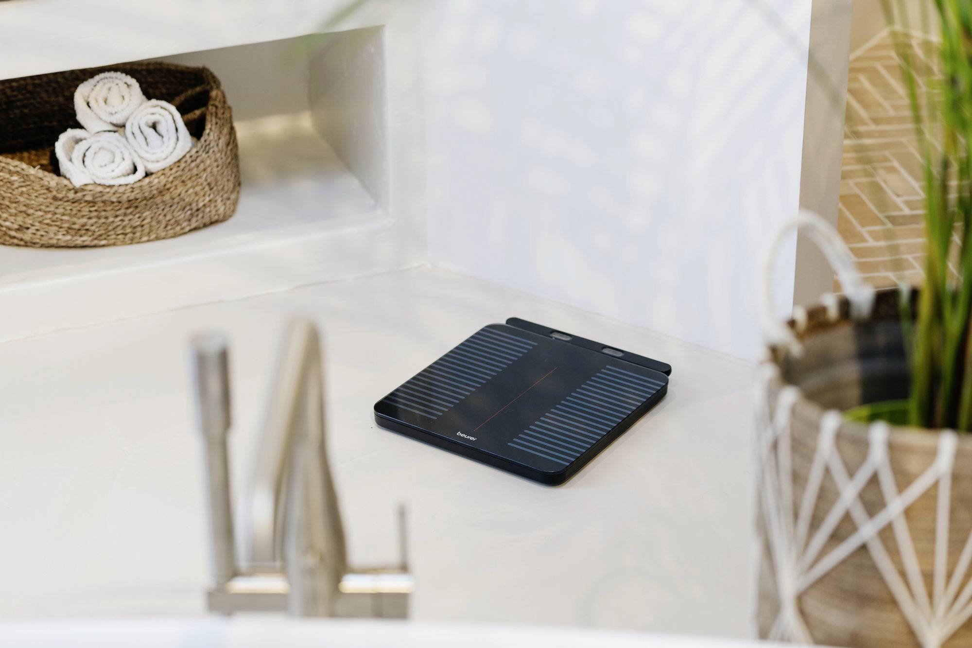 Eine Personenwaage auf einem weißen Boden neben einer Pflanze und einem Korb mit zusammengerollten Handtüchern in einer modernen Badezimmereinrichtung.