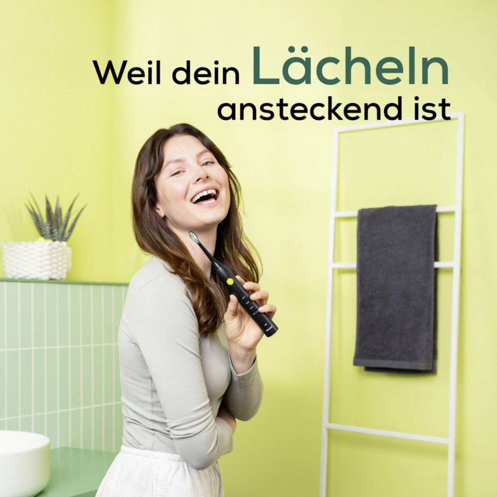 A woman in a bathroom smiles while holding an electric toothbrush. The text reads 'Weil dein Lächeln ansteckend ist' (Because your smile is contagious).(Note: The text is already in German, so no translation is needed.)