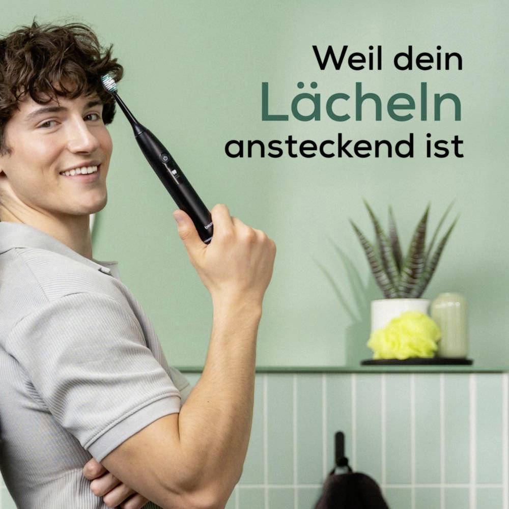 Here's the translation to German:Ein Person, die Zähne putzt und lächelt, neben dem Text 'Weil dein Lächeln ansteckend ist' in einem Badezimmer mit Topfpflanzen.