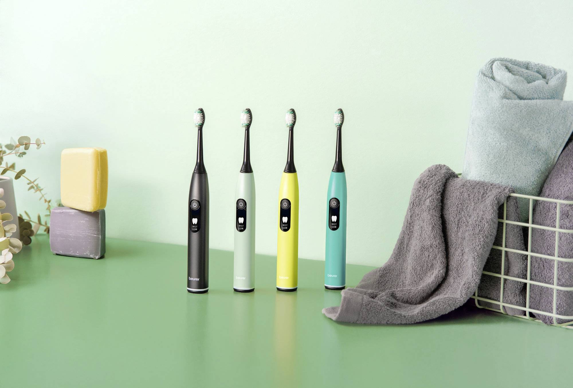 Vier elektrische Zahnbürsten in verschiedenen Farben stehen aufrecht auf einer mintgrünen Oberfläche neben zusammengelegten Handtüchern und Seifenstücken.