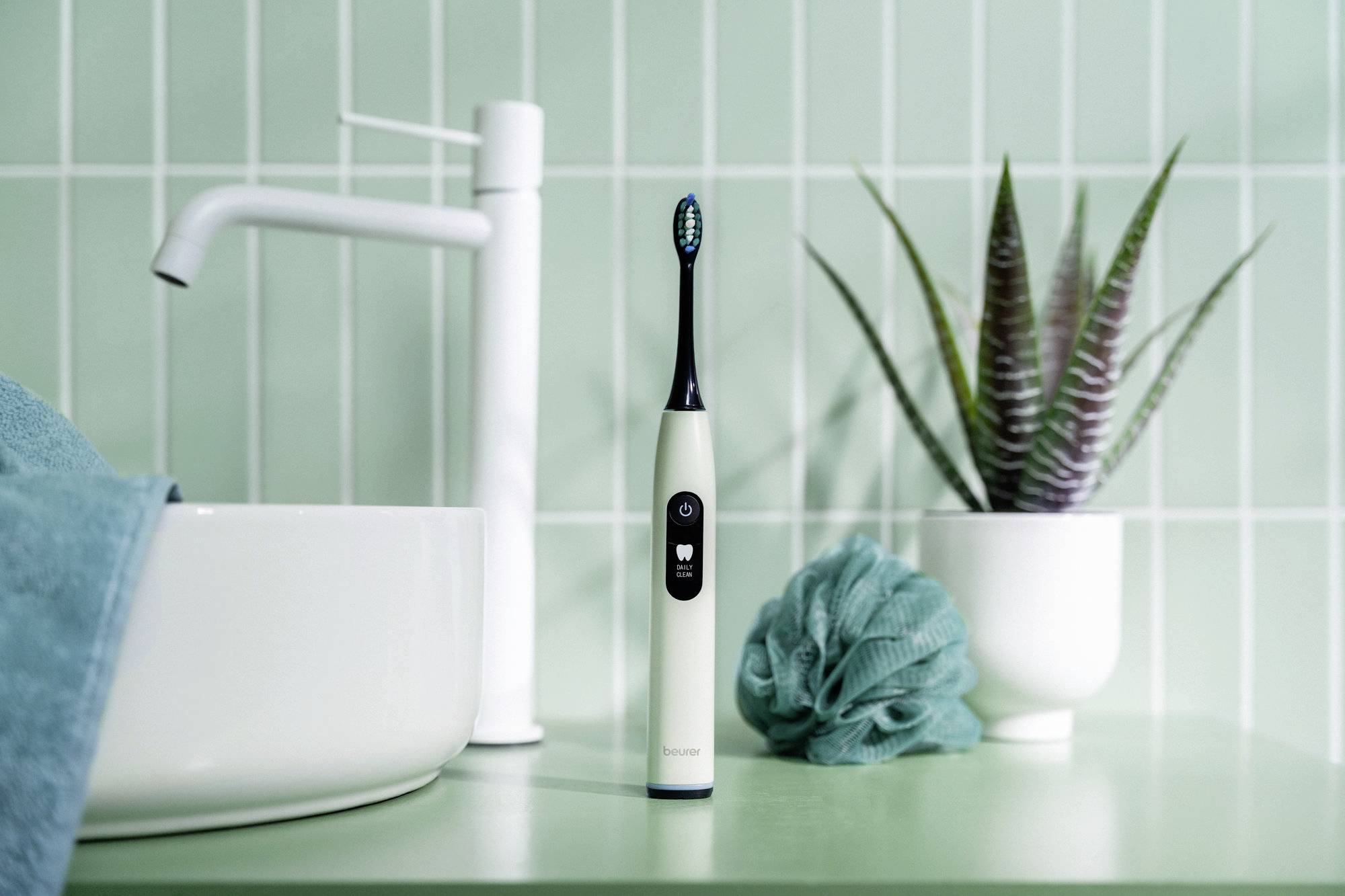 Elektrische Zahnbürste steht auf grüner Badezimmerablage neben einem modernen weißen Wasserhahn, Handtuch, grünem Badeschwamm und Topfpflanze.